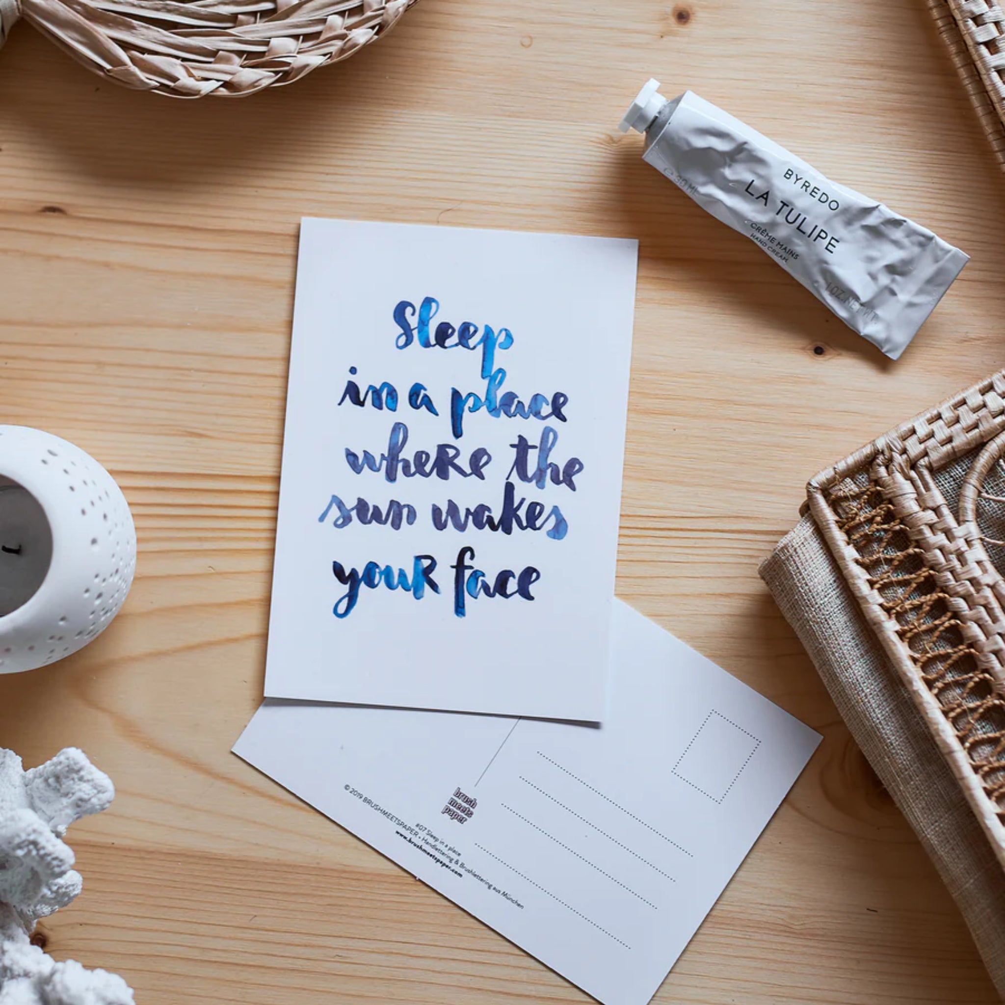 Karte mit dem Zitat "Sleep in a place where the sun wakes your face" auf einem Holztisch neben einer Tube Creme und Kerze.