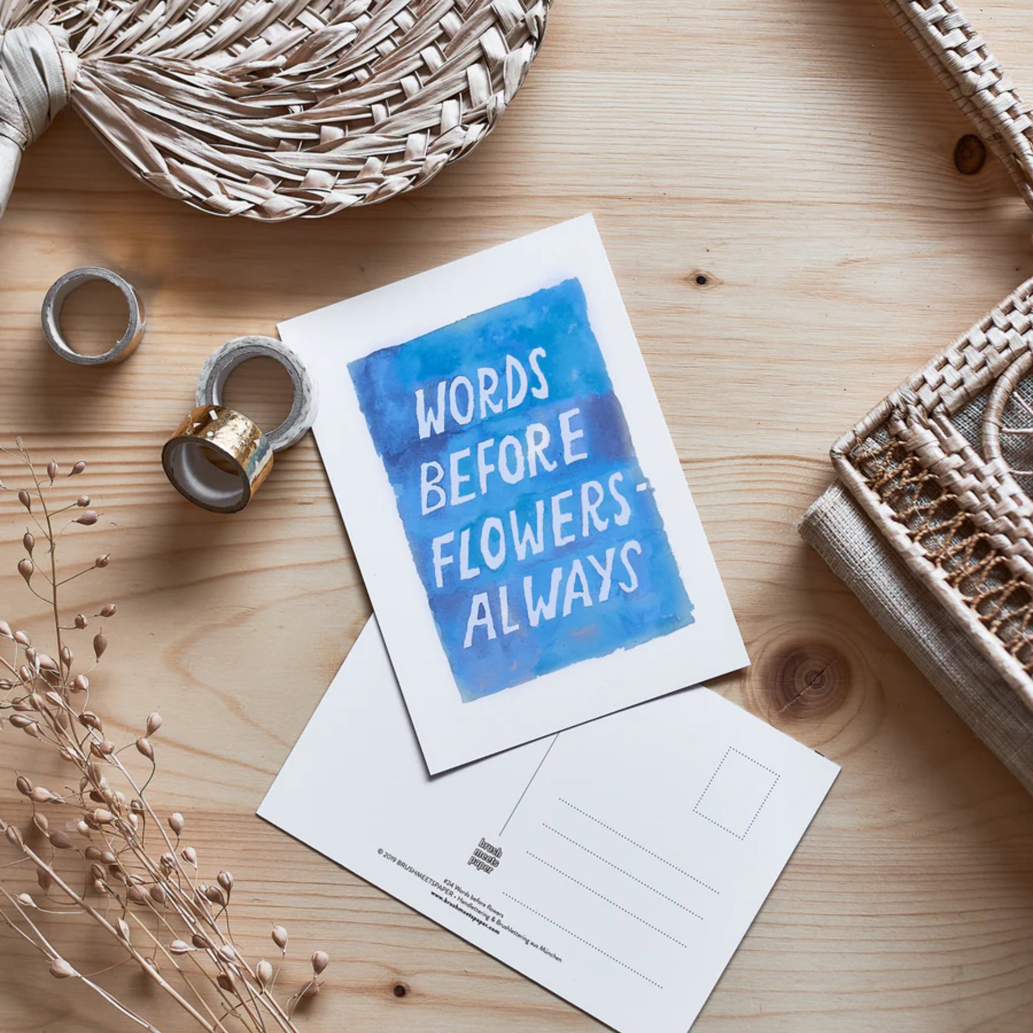 Postkarte auf Holzuntergrund mit dem Text "WORDS BEFORE FLOWERS ALWAYS" in blauer Schrift, umgeben von Bastelmaterialien.