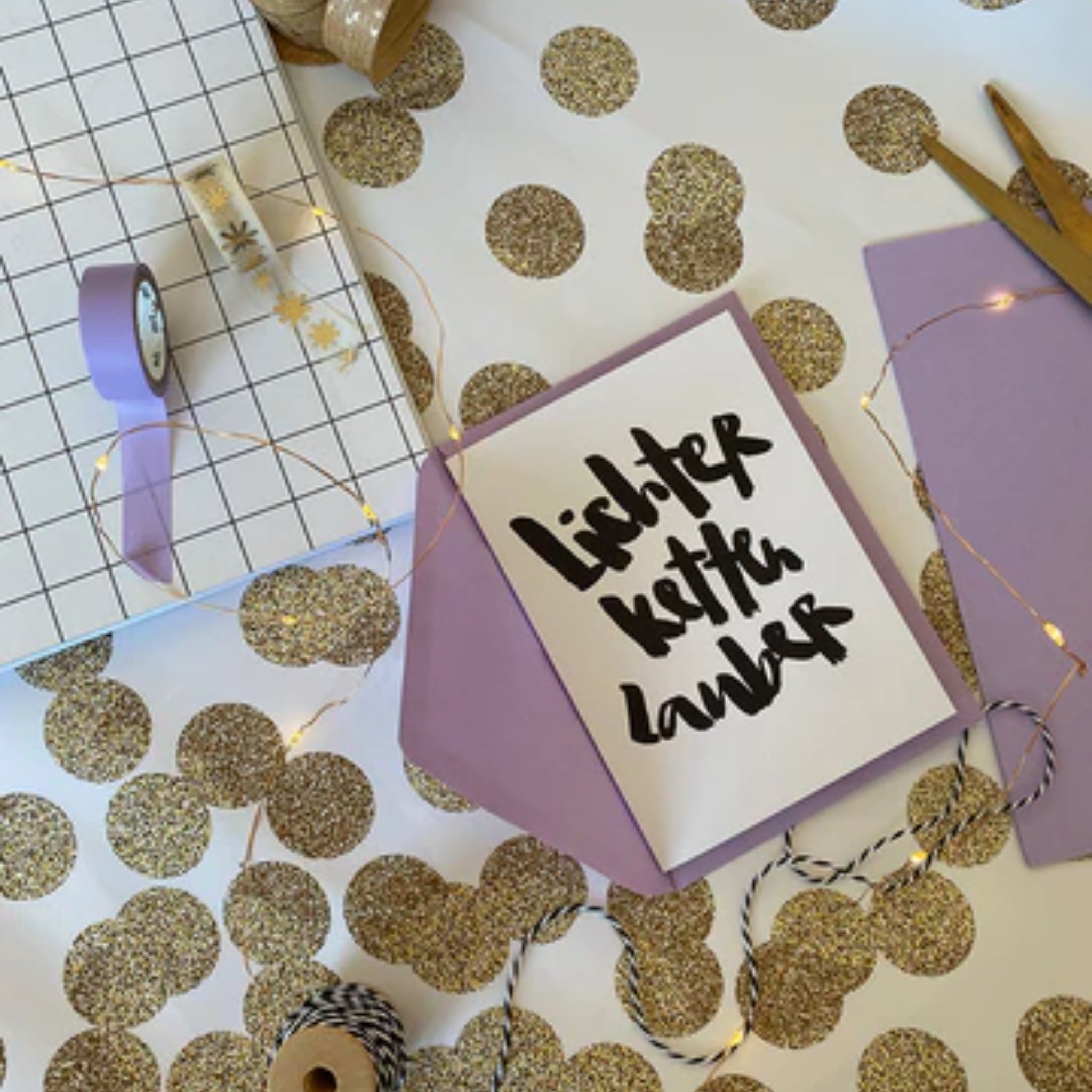 Bastelmaterialien und eine Karte mit „Lichterkette Zauber“ auf einem Tisch mit glitzernden Punkten und Lichterkette.