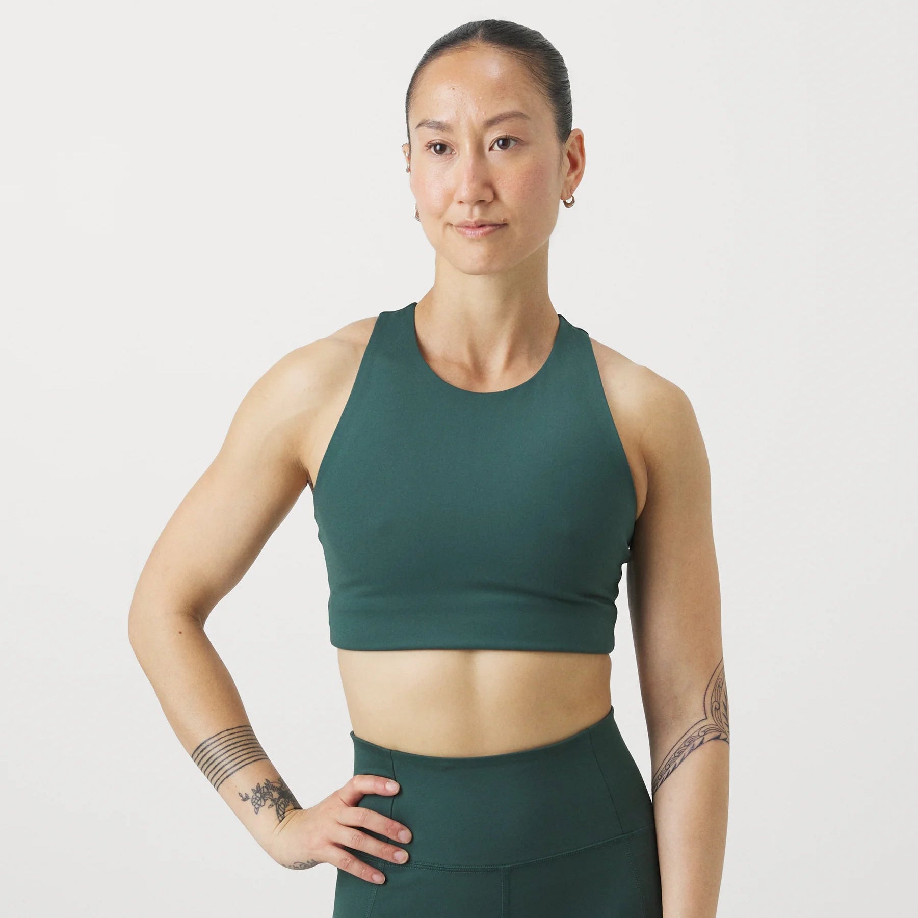 Eine Person, die ein dunkelgrünes, sportliches Crop-Top und passende Leggings trägt, steht vor einem weißen Hintergrund mit einer Hand an der Hüfte und blickt selbstbewusst zur Seite. Sie hat Tätowierungen auf den Armen und ihr Haar ist zurückgebunden.