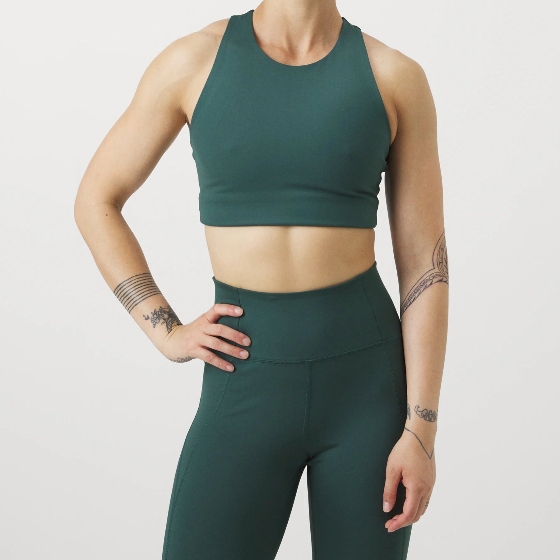 Eine Frau in einem dunkelgrünen Sport-BH und Leggings steht vor einem weißen Hintergrund und stützt eine Hand auf ihre Hüfte. Sie hat Tattoos auf beiden Armen und trägt ihr Haar zu einem ordentlichen Dutt.
