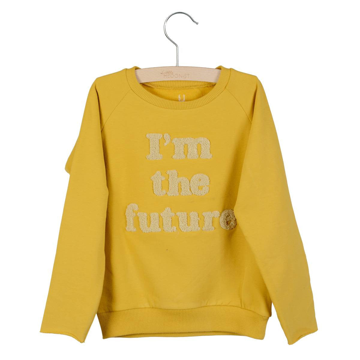 Ein gelbes, langärmeliges Sweatshirt auf einem Bügel mit der Aufschrift Im the future in strukturierten Buchstaben auf der Vorderseite.