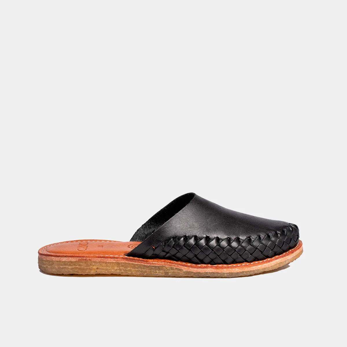 Ein schwarzer Slipper aus Leder mit einem gewebten Seitenteil, einer hellbraunen Lederinnensohle und einer flachen, strukturierten braunen Sohle, im Profil auf einem einfarbig weißen Hintergrund dargestellt.