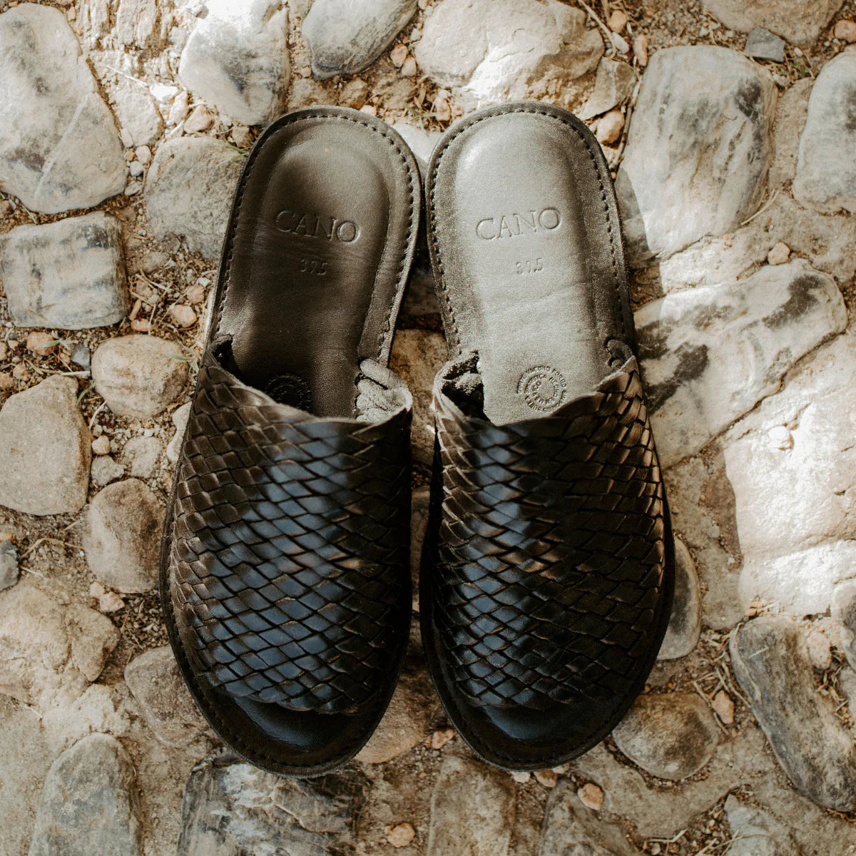 Ein Paar Slipper aus gewebtem Leder mit dem eingeprägten Markennamen CANO auf der Innensohle, die nebeneinander auf einem Kopfsteinpflaster stehen.