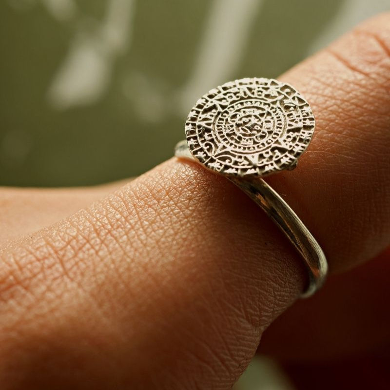 Nahaufnahme eines Silberrings mit einem komplizierten, antiken, münzähnlichen Design am Finger einer Person, mit detaillierten Mustern und Symbolen auf der kreisförmigen Oberfläche des Rings.