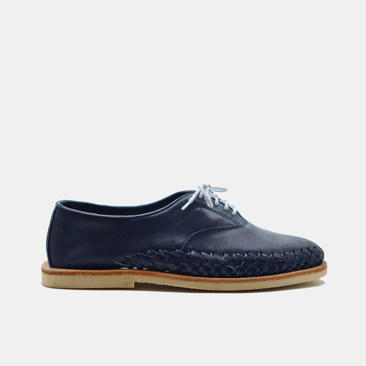 Ein marineblauer Leder-Oxford-Schuh mit weißen Schnürsenkeln, einer hellbraunen Kreppsohle und einem markanten gewebten Lederdetail an der Seite, das vor einem schlichten weißen Hintergrund steht.