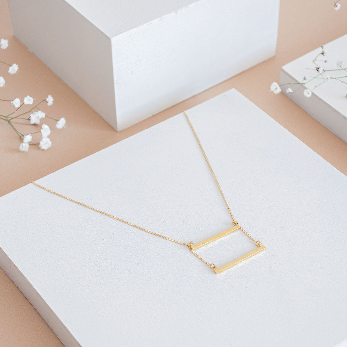 Eine zarte goldene Halskette mit einem rechteckigen Stabanhänger steht auf einer weißen rechteckigen Plattform, umgeben von weißen geometrischen Blöcken und kleinen Zweigen mit Schleierkrautblüten auf einem beigen Hintergrund.