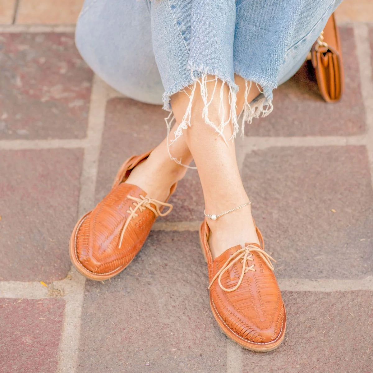 Eine Person mit hellbraunen Schuhen aus gewebtem Leder und einer blauen Jeans mit ausgefranstem Saum sitzt auf einer gefliesten Fläche und hat die Beine an den Knöcheln gekreuzt. Ein Knöchel ist mit einem kleinen Fußkettchen geschmückt.