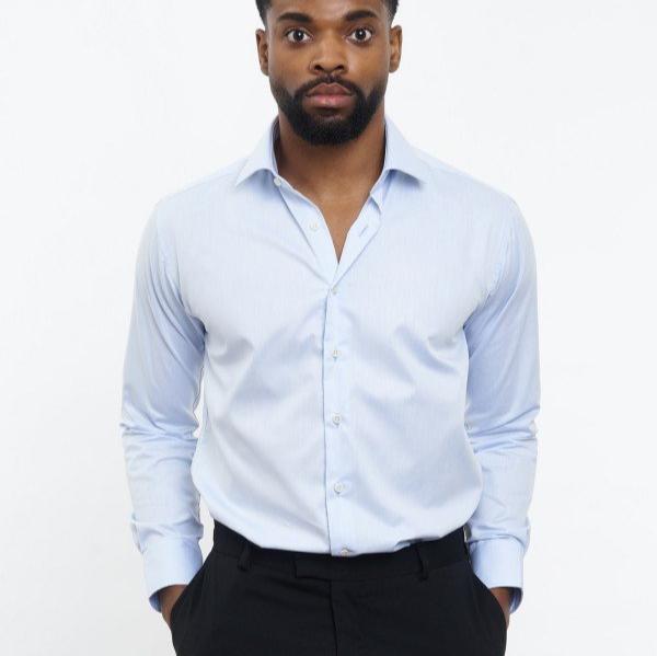 Ein Mann in einem hellblauen Hemd und dunkler Hose mit den Händen in den Taschen vor weißem Hintergrund.