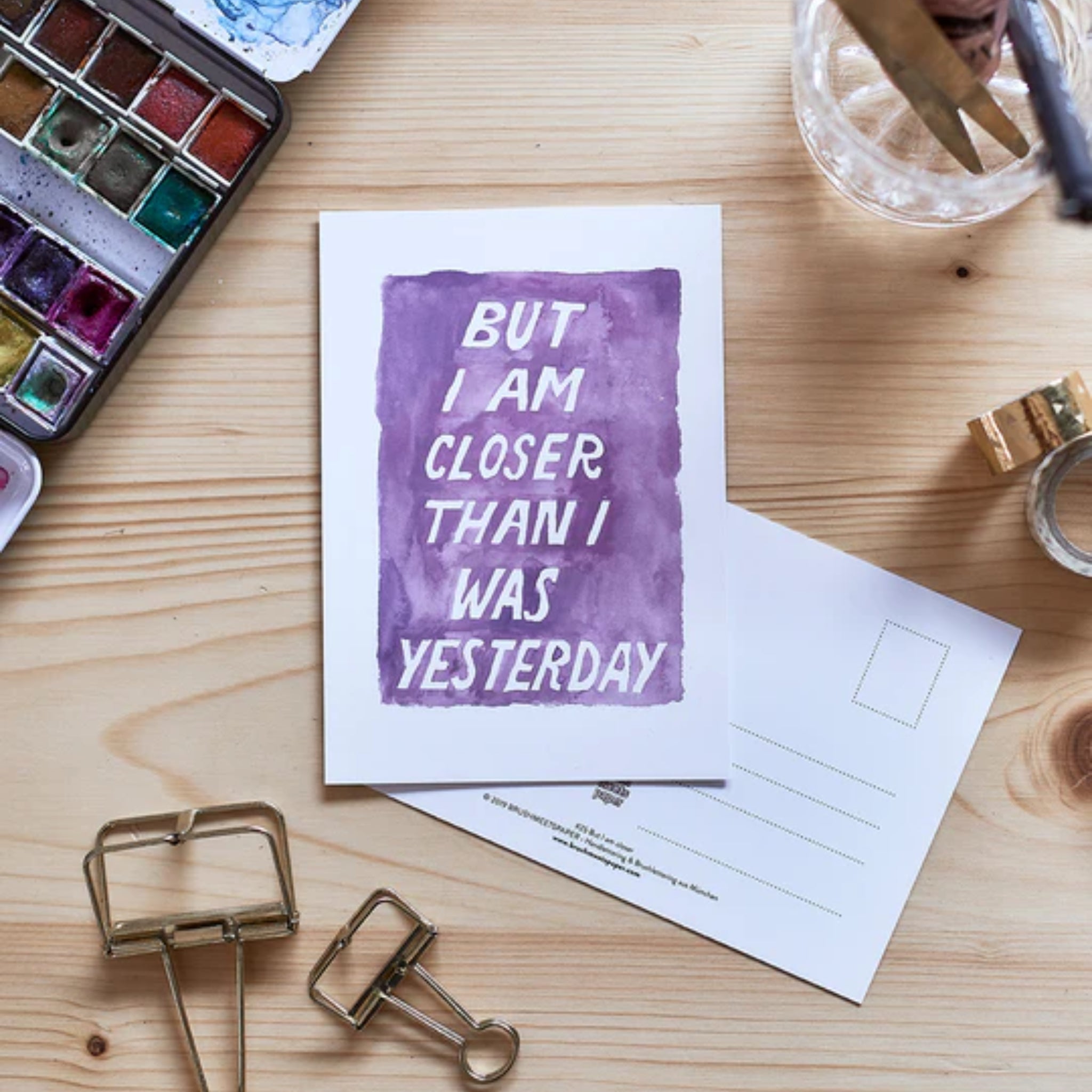 Eine Karte mit lilafarbenem Text "BUT I AM CLOSER THAN I WAS YESTERDAY" auf einem Holztisch mit Kunstmaterialien.