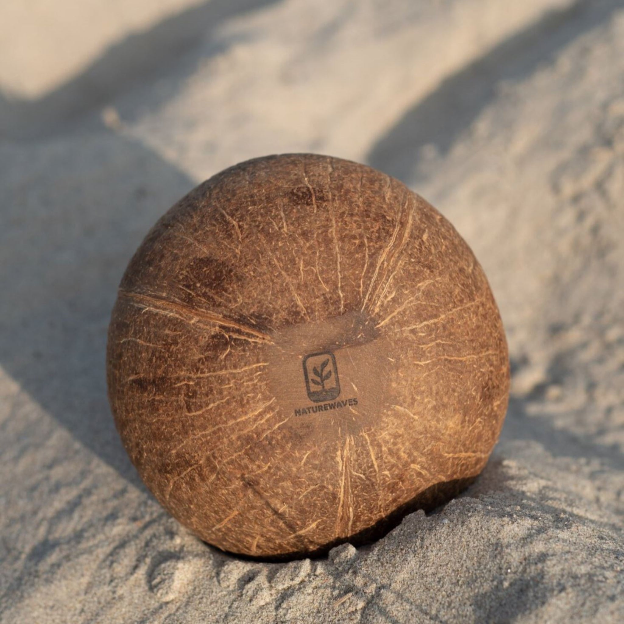 Eine braune Kokosnuss mit dem Logo "Naturewaves" liegt auf hellem Sand.