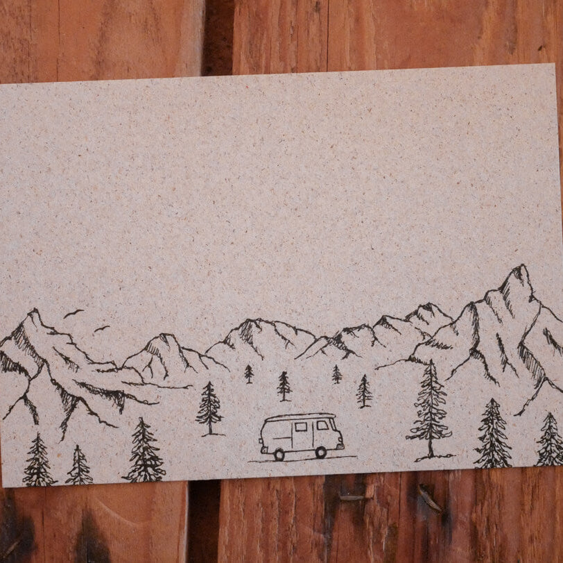 Eine einfache schwarze Tuschezeichnung auf braunem Papier zeigt Berge, Kiefern und einen Lieferwagen in einer Waldlandschaft. Das Papier liegt auf einer Holzunterlage.