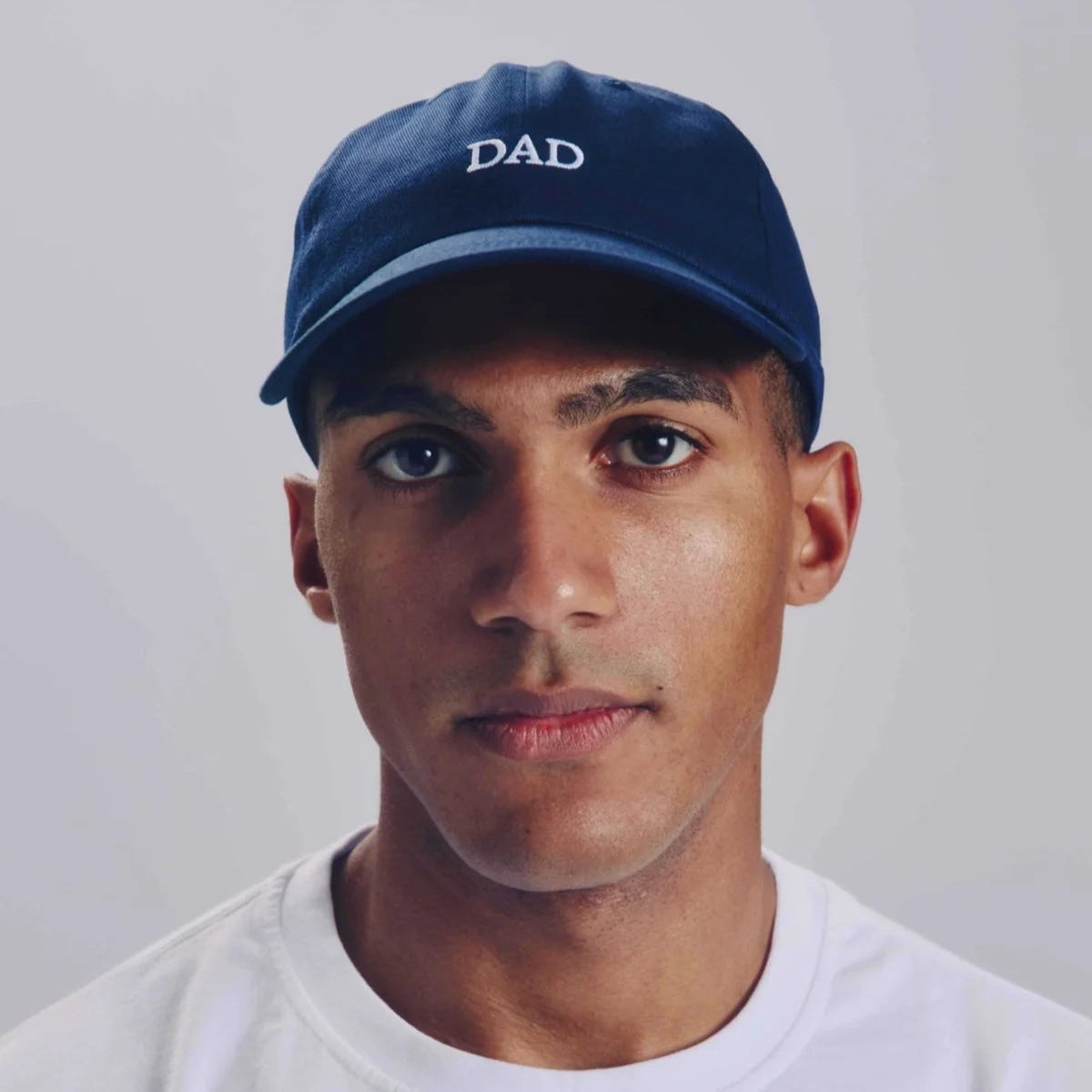 Eine Person, die eine marineblaue Baseballkappe mit dem aufgestickten Wort DAD und ein schlichtes weißes T-Shirt trägt, blickt vor einem hellgrauen Hintergrund in die Kamera.