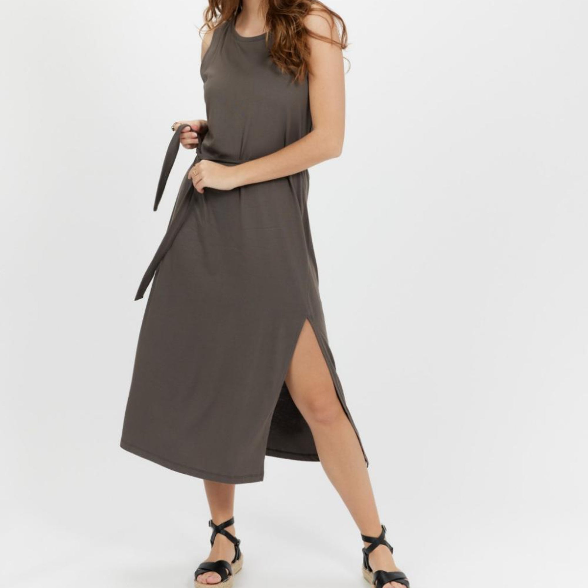 Frau in einem ärmellosen, grauen Kleid mit Seitenschlitz, das mit einem Gürtel gebunden ist, trägt schwarze Sandalen.