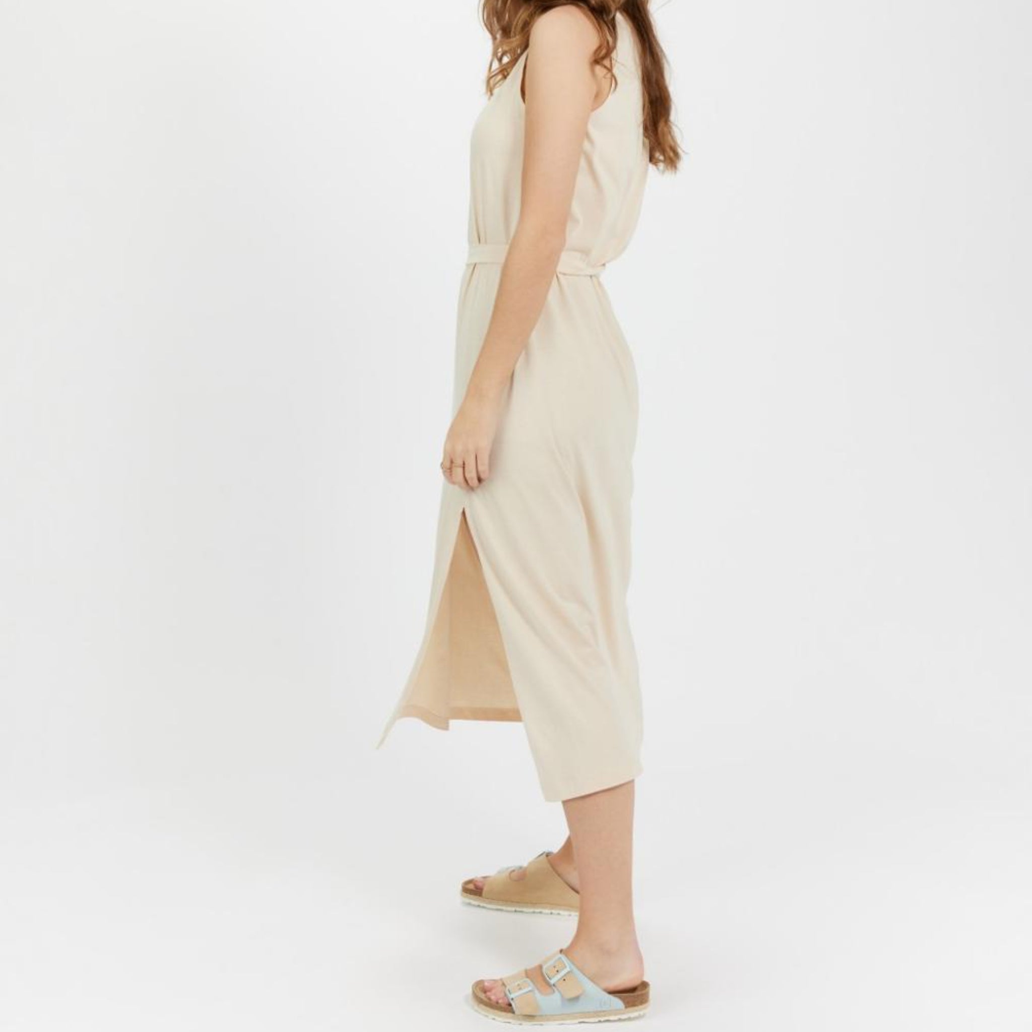 Person in a beige, sleeveless dress with a side slit, wearing sandals, stands against a plain white background.