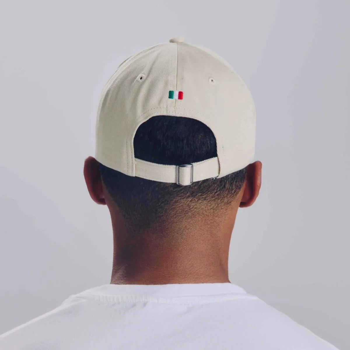 Eine Person, die eine weiße Mütze und ein weißes T-Shirt trägt, wird von hinten gezeigt. Auf der Mütze ist oberhalb des verstellbaren Riemens eine kleine mexikanische Flagge aufgestickt. Der Hintergrund ist einfarbig und hell.