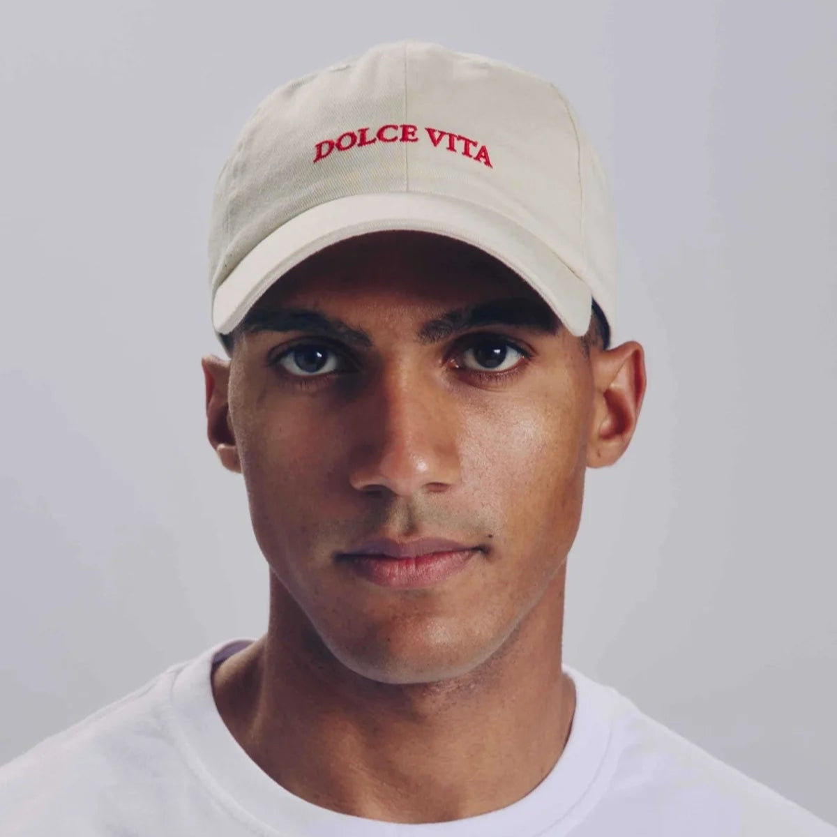 Eine Person, die eine beigefarbene Kappe mit rotem DOLCE VITA-Schriftzug und ein schlichtes weißes T-Shirt trägt, blickt nach vorne auf einen hellgrauen Hintergrund.