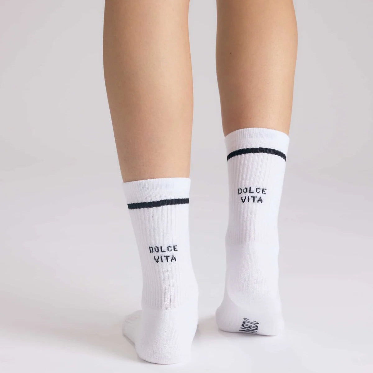 Eine Person steht auf einer weißen Fläche und trägt weiße Ringelsocken mit einem schwarzen Streifen am oberen Ende und der Aufschrift DOLCE VITA auf der Rückseite jeder Socke. Die Socken reichen bis zur Mitte der Wade.