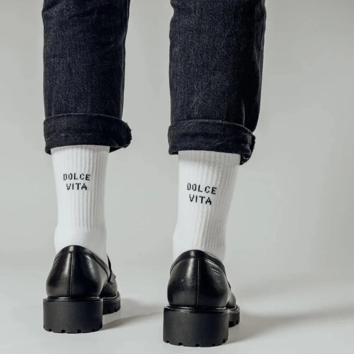 Eine Person, die schwarze Schuhe, dunkle, hochgekrempelte Jeans und weiße Socken mit der Aufschrift DOLCE VITA trägt, steht vor einem schlichten hellen Hintergrund.