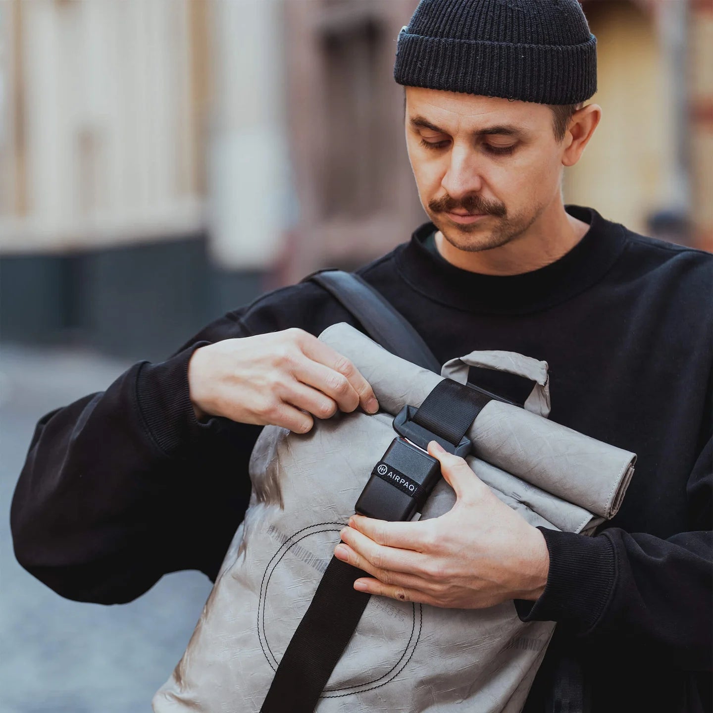 Ein Mann mit schwarzer Mütze und Sweatshirt schließt die Schnalle eines hellgrauen Rolltop-Rucksacks, während er auf einer Straße in der Stadt steht.