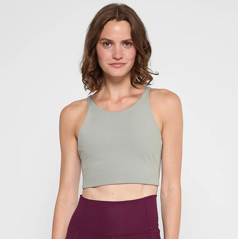 Eine Frau mit gewelltem braunem Haar, die ein hellgraues ärmelloses Crop-Top und kastanienbraune Leggings mit hoher Taille trägt, steht vor einem schlichten weißen Hintergrund und lächelt sanft in die Kamera.