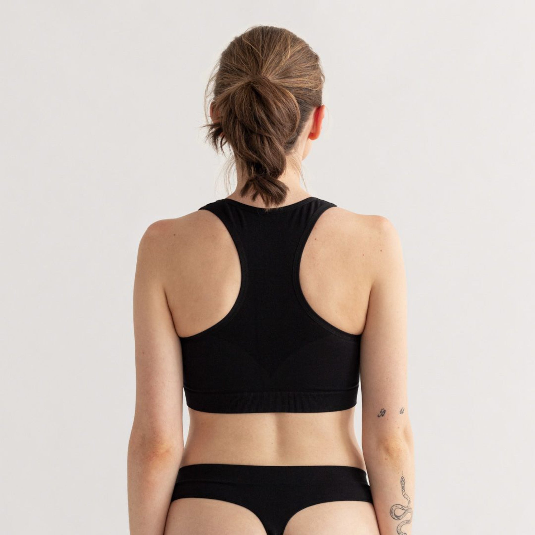 Frau in schwarzer Sportbekleidung von hinten, mit einem Tattoo auf dem Arm, vor einem neutralen Hintergrund.