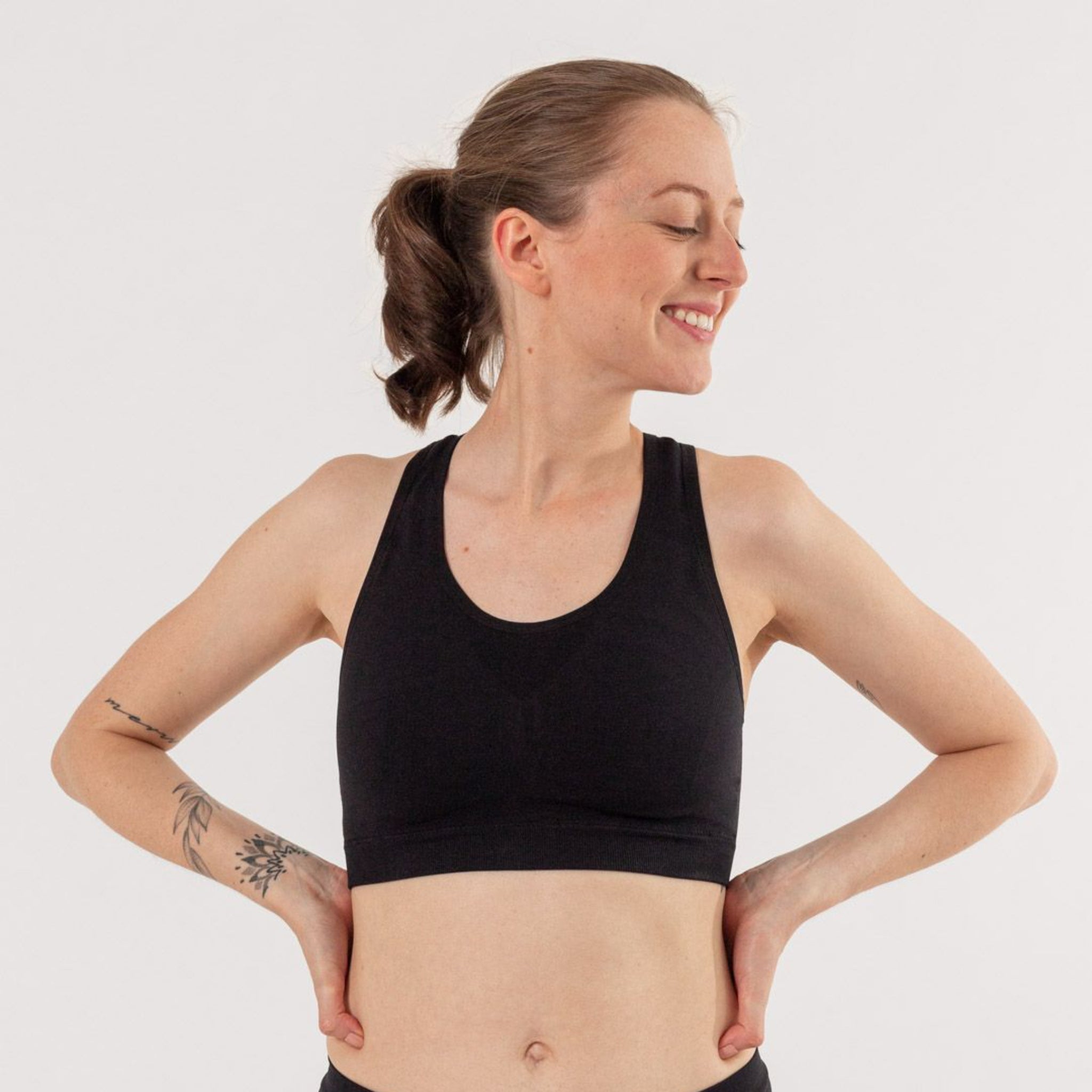 Lächelnde Frau in Sport-BH posiert mit geschlossenen Augen vor weißem Hintergrund, Tätowierungen an den Armen.