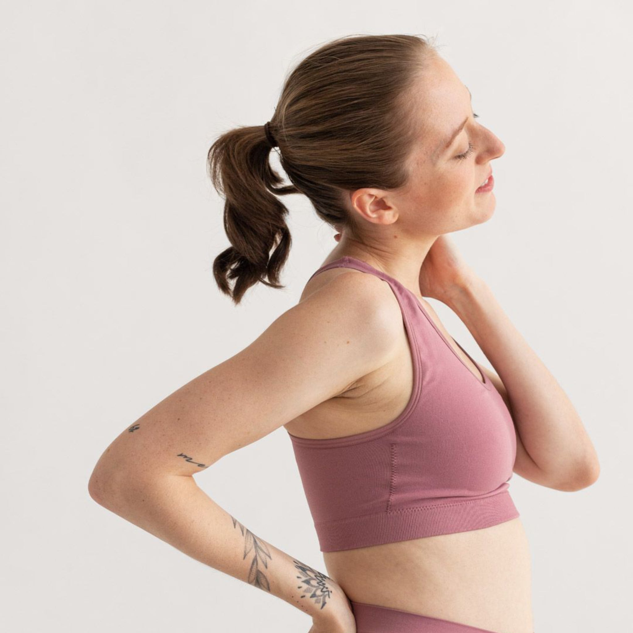 Frau in sportlichem rosa Top fasst sich an den Nacken und scheint zu strecken, mit tätowiertem Arm sichtbar.