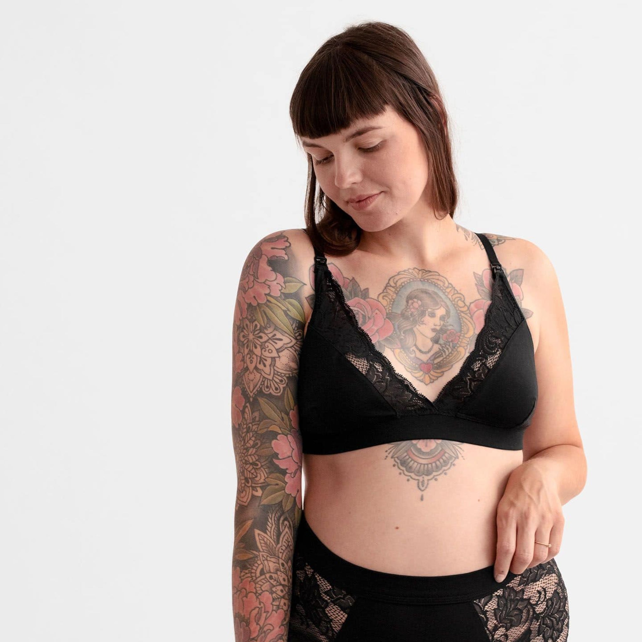 Frau mit blumigen Tattoos trägt schwarze Spitzenunterwäsche und blickt zur Seite, vor weißem Hintergrund.