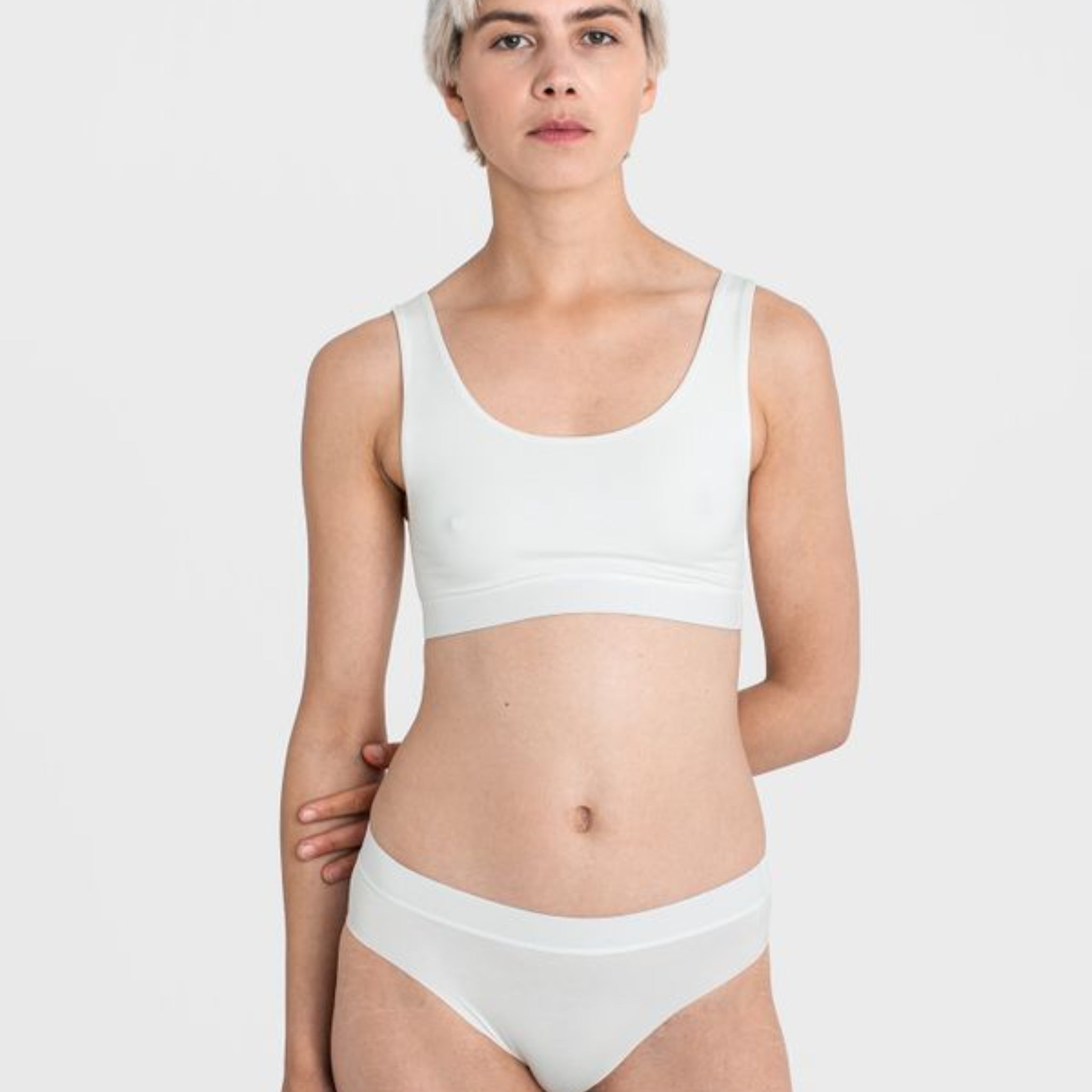 Eine Person steht in weißer Unterwäsche vor neutralem Hintergrund.