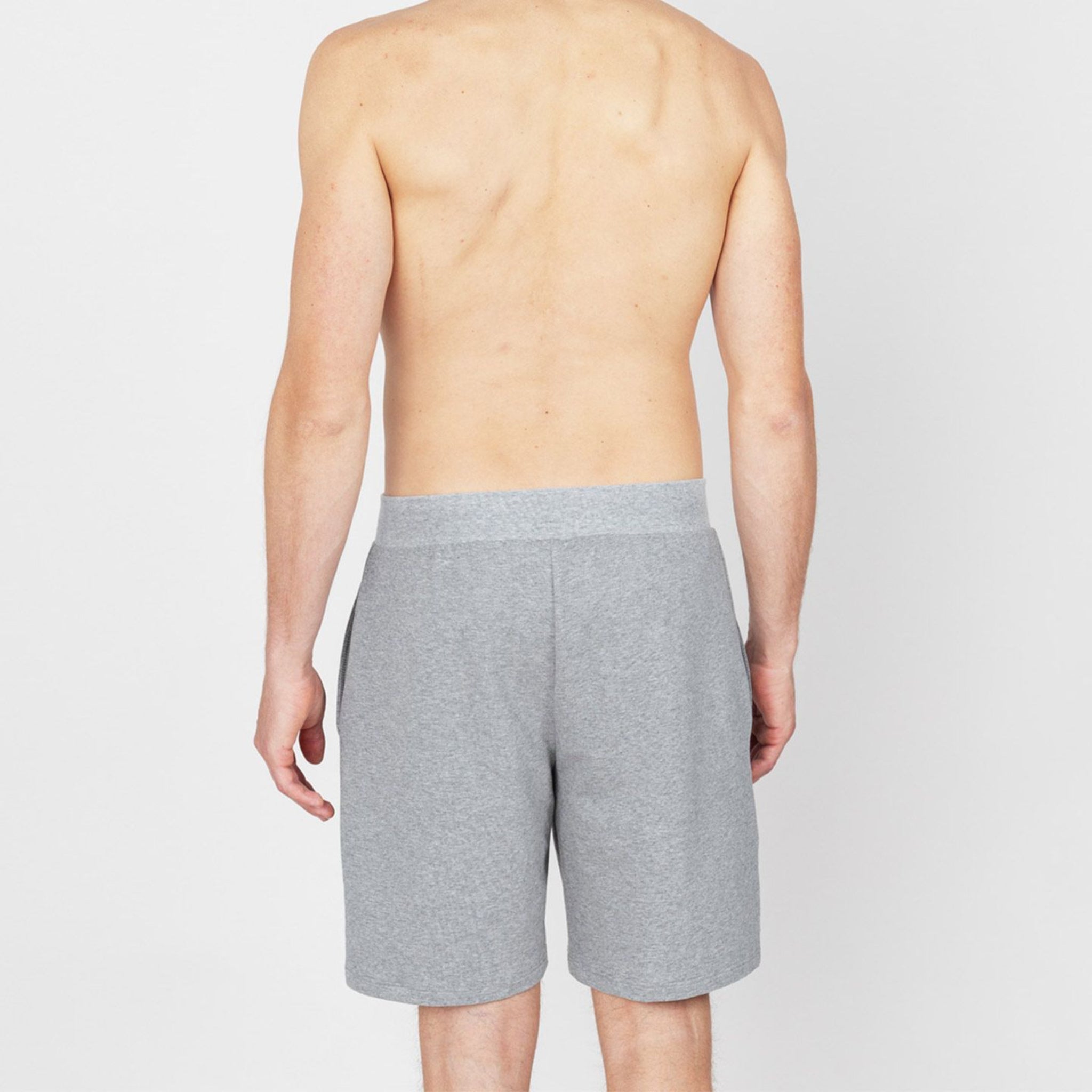 Ein Mann ohne Shirt steht mit grauen Shorts mit dem Rücken zur Kamera vor einem weißen Hintergrund.