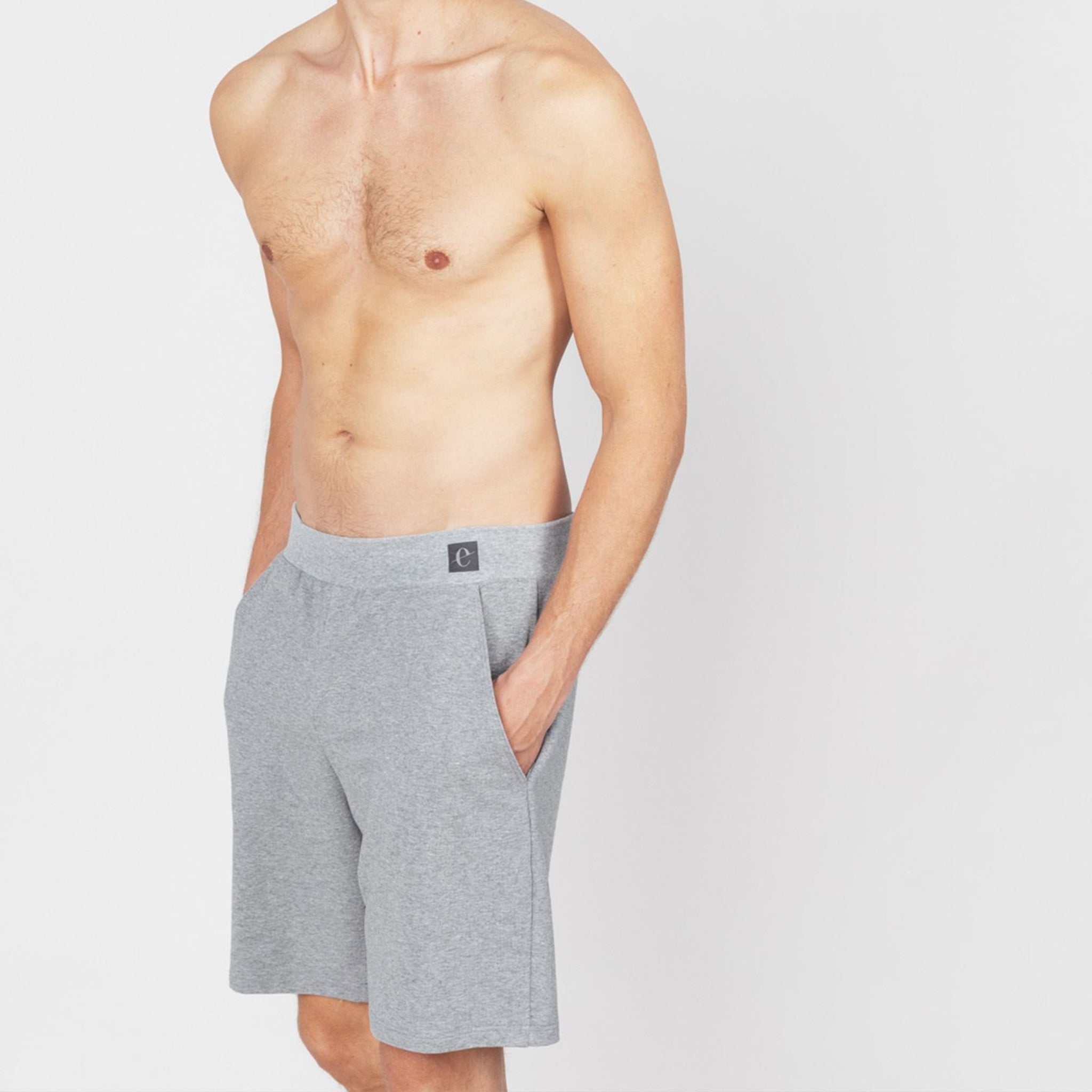 Ein unbekleideter Mann steht, trägt graue Shorts und hat die Hände in den Taschen.