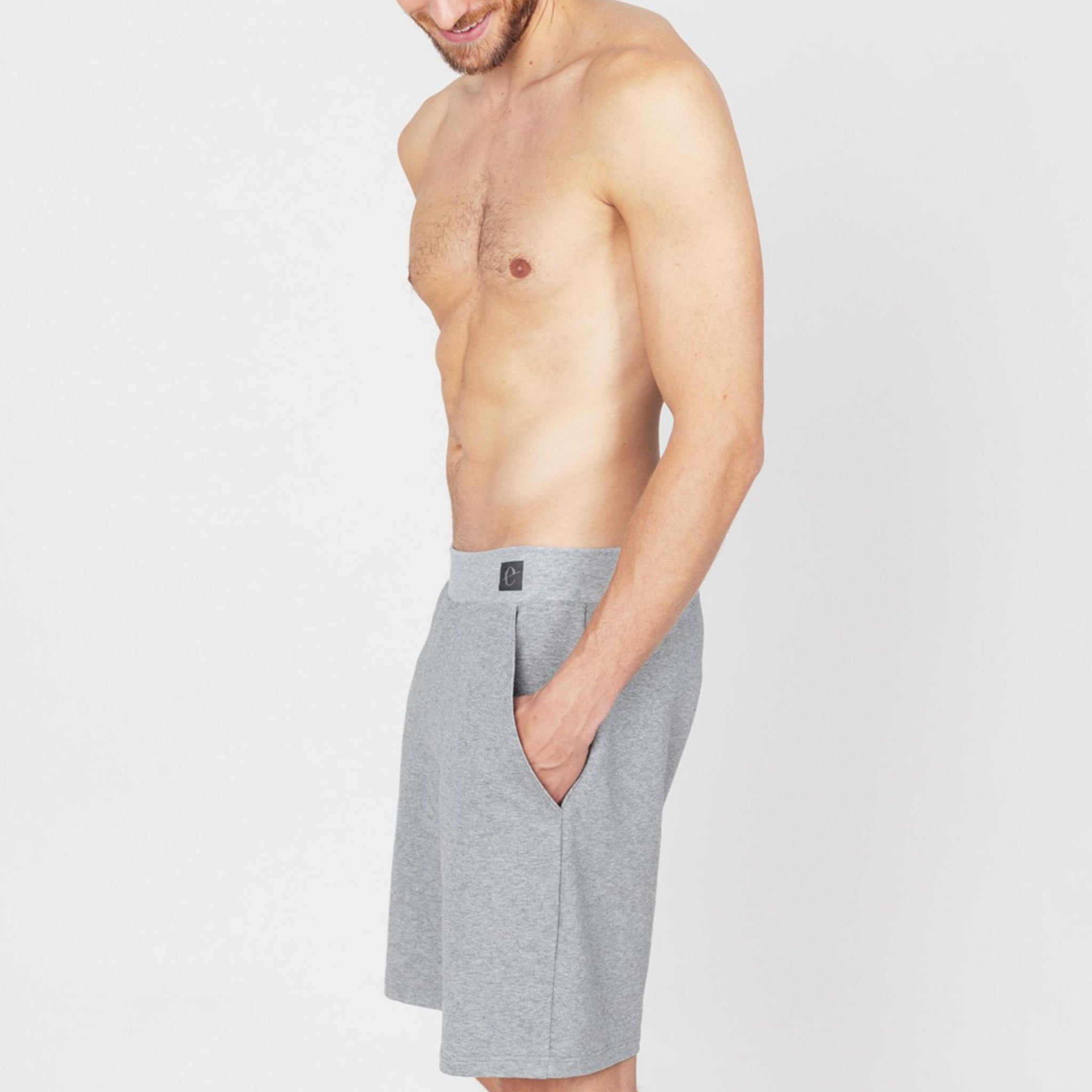 Oberkörperfreier Mann in grauen Shorts steht seitlich vor einem weißen Hintergrund.