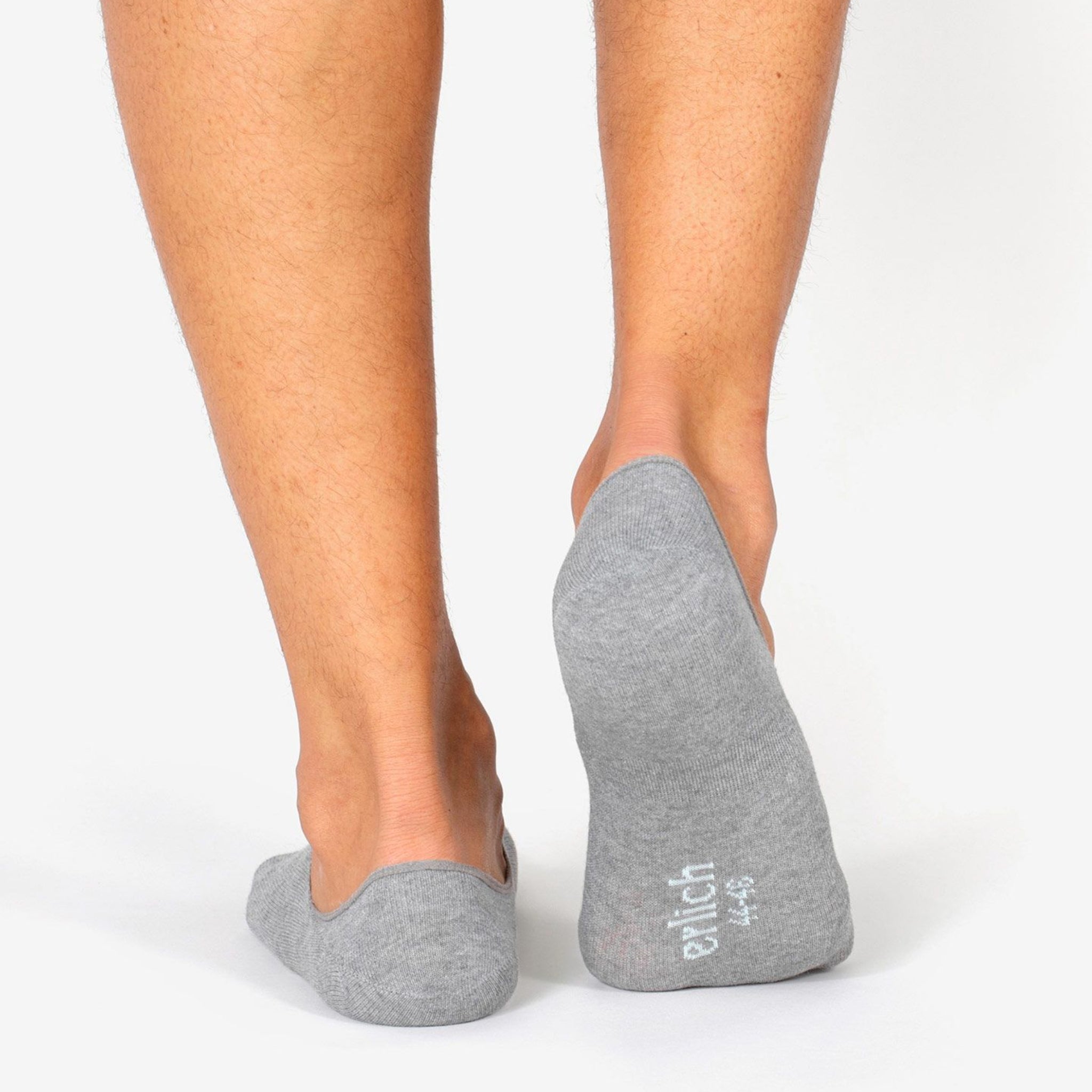 Rückansicht von Beinen mit grauen Füßlingen, der linke Fuß gehoben, Markenname auf der Sohle des rechten Fußes.