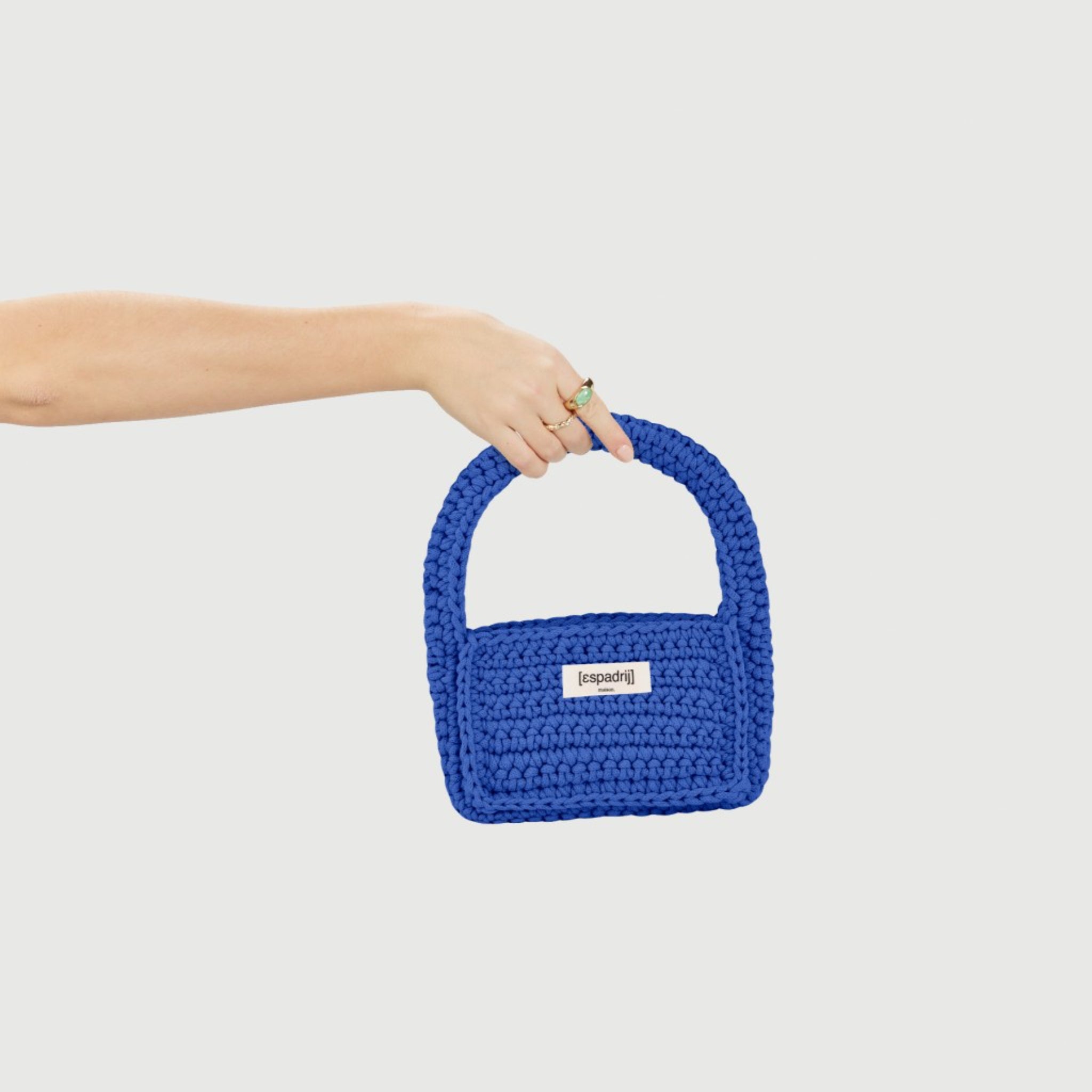 Eine Hand hält eine blaue, handgefertigte Tasche mit kurzem Henkel auf einem weißen Hintergrund.