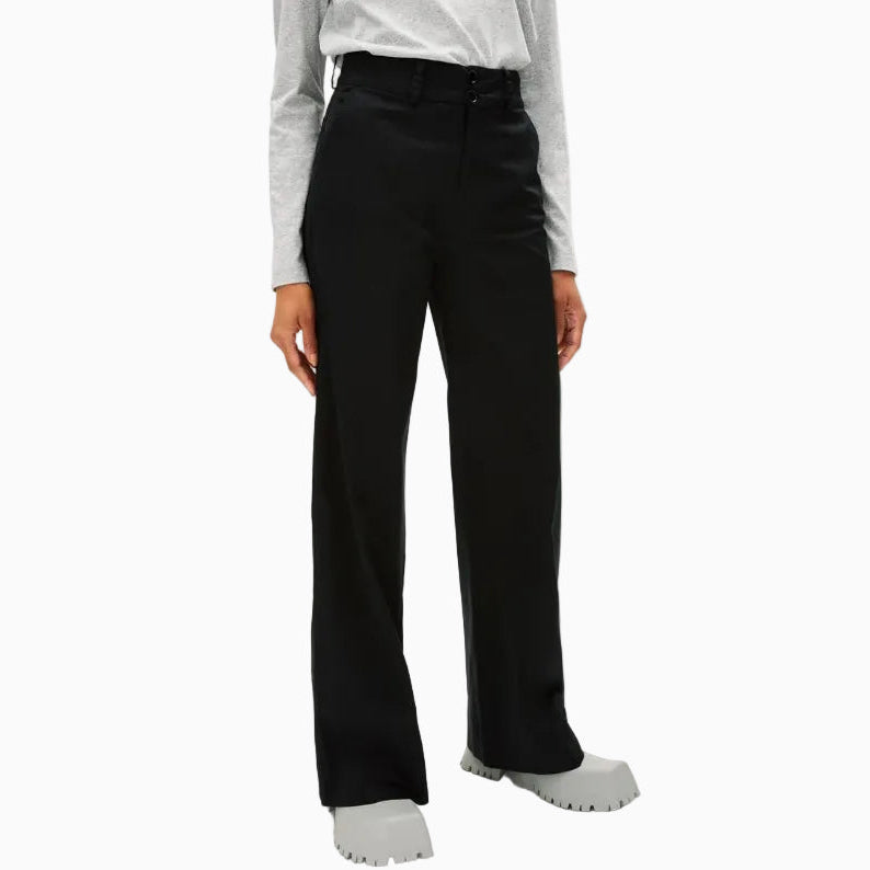 Eine Person, die ein hellgraues langärmeliges Oberteil, eine schwarze Hose mit weitem Bein und graue Schuhe mit klobigen Sohlen trägt, steht vor einem einfarbig weißen Hintergrund.