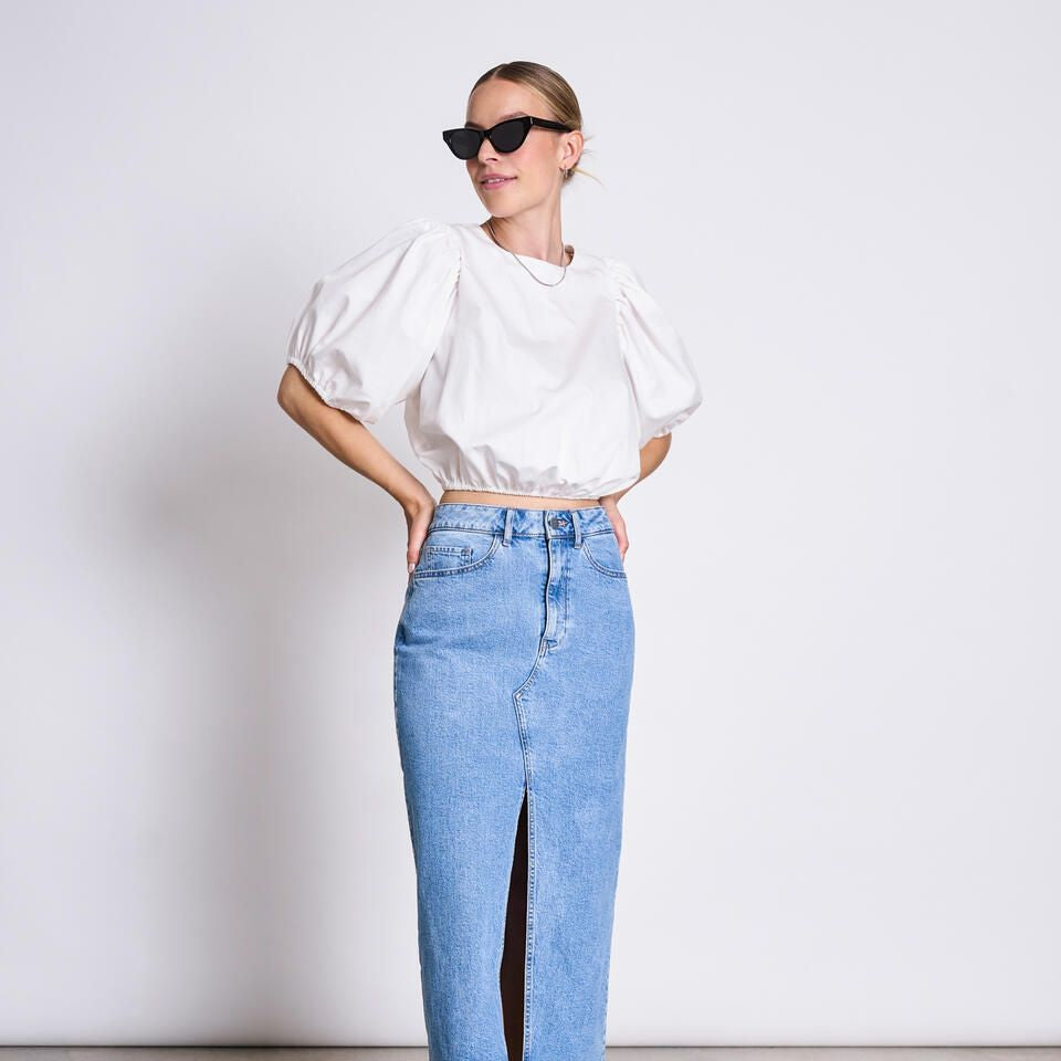 Frau in weißer Bluse und langer Jeansrock mit Schlitz, trägt Sonnenbrille, vor weißem Hintergrund.