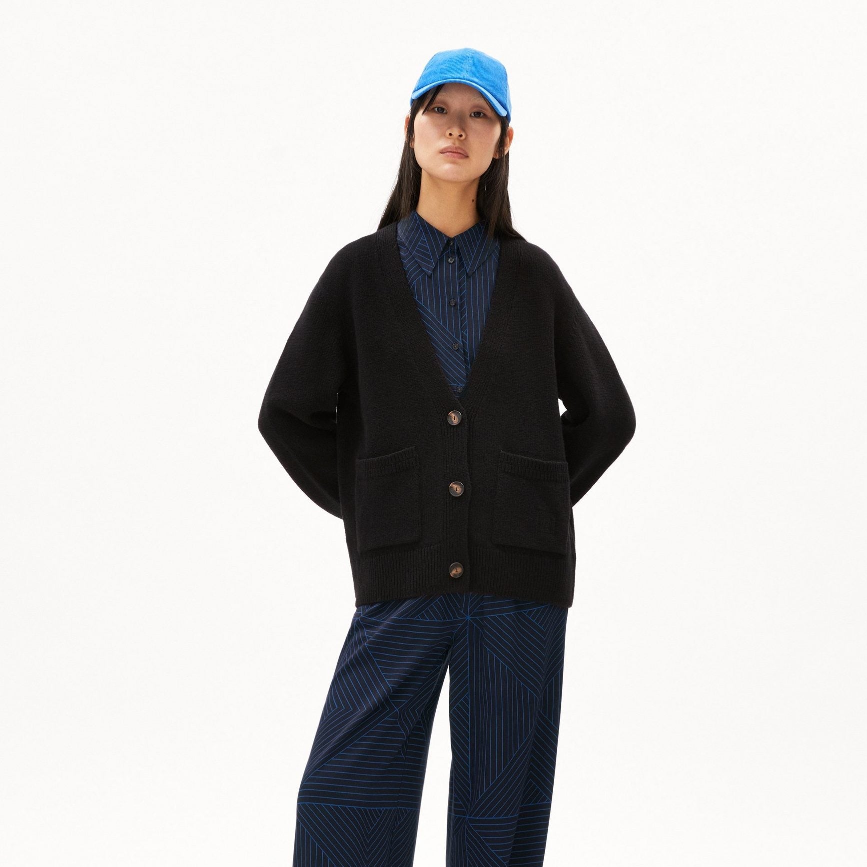 Eine Frau steht mit den Händen auf dem Rücken, trägt eine schwarze Strickjacke über einem blau gemusterten Hemd und eine passende Hose mit weitem Bein. Außerdem trägt sie eine hellblaue Kappe und blaue Turnschuhe vor einem schlichten weißen Hintergrund.