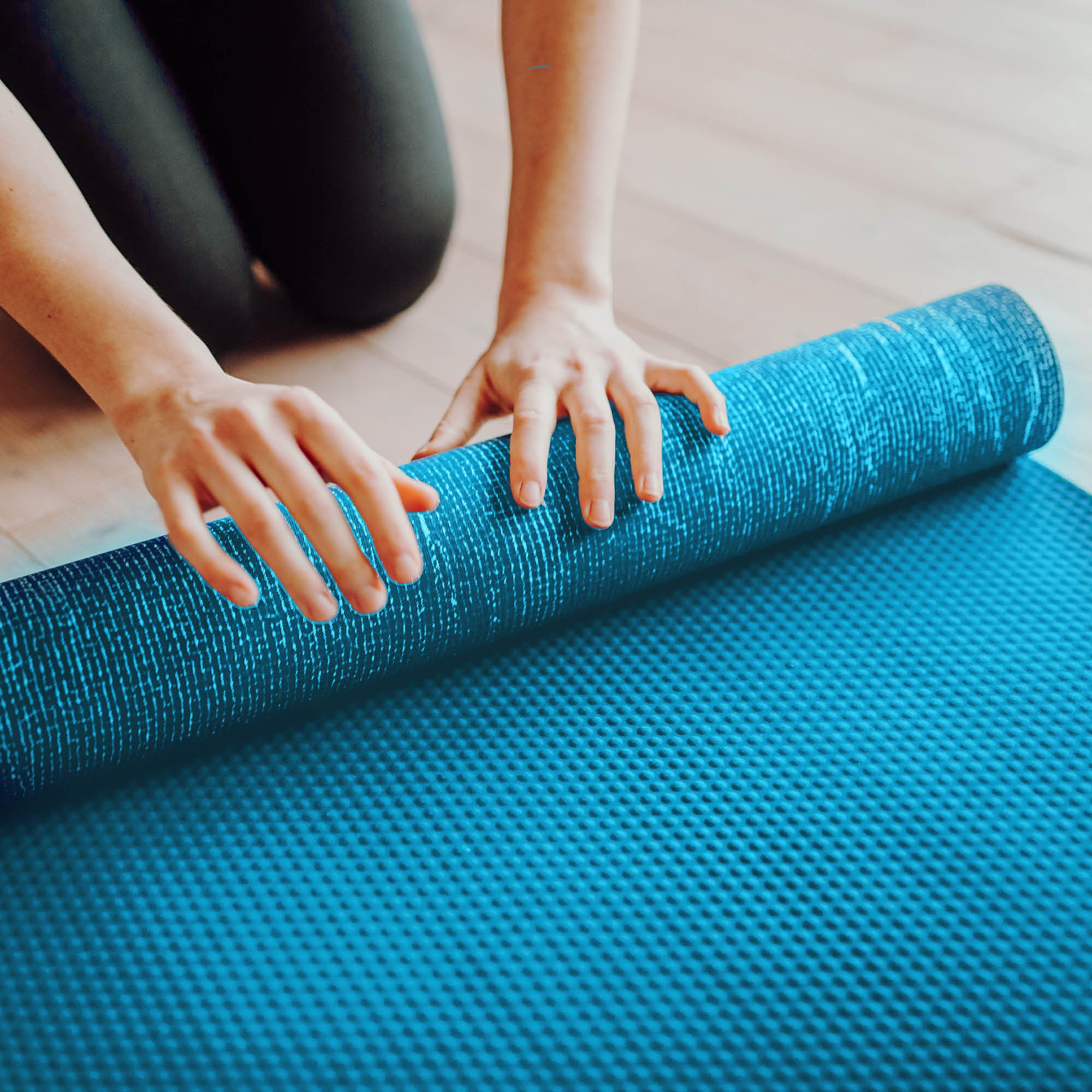 Eine Person kniet auf einem Holzboden und rollt mit beiden Händen eine blaue, strukturierte Yogamatte auf.