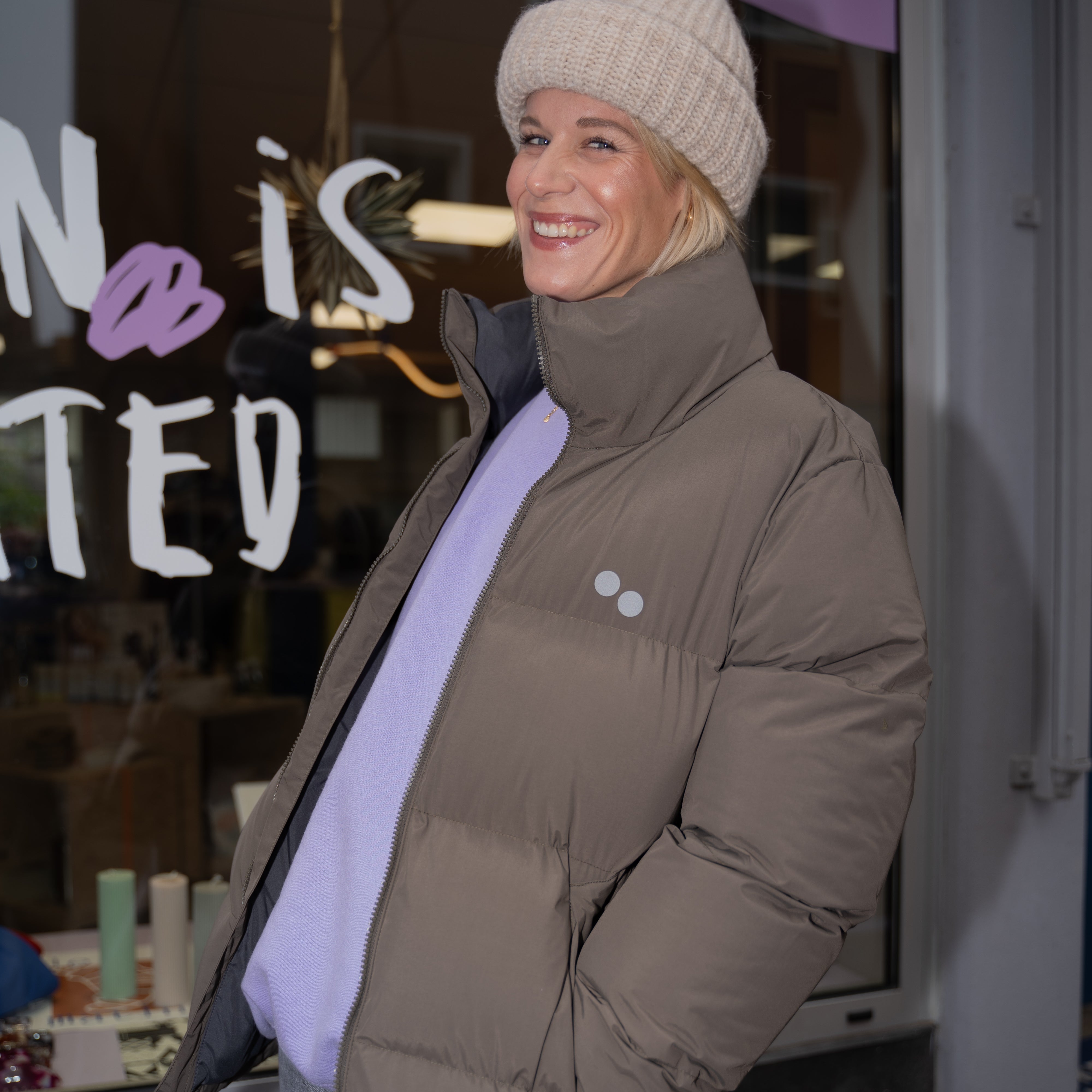 Frau in einer beigen Mütze und brauner Jacke lächelt vor einem Schaufenster.