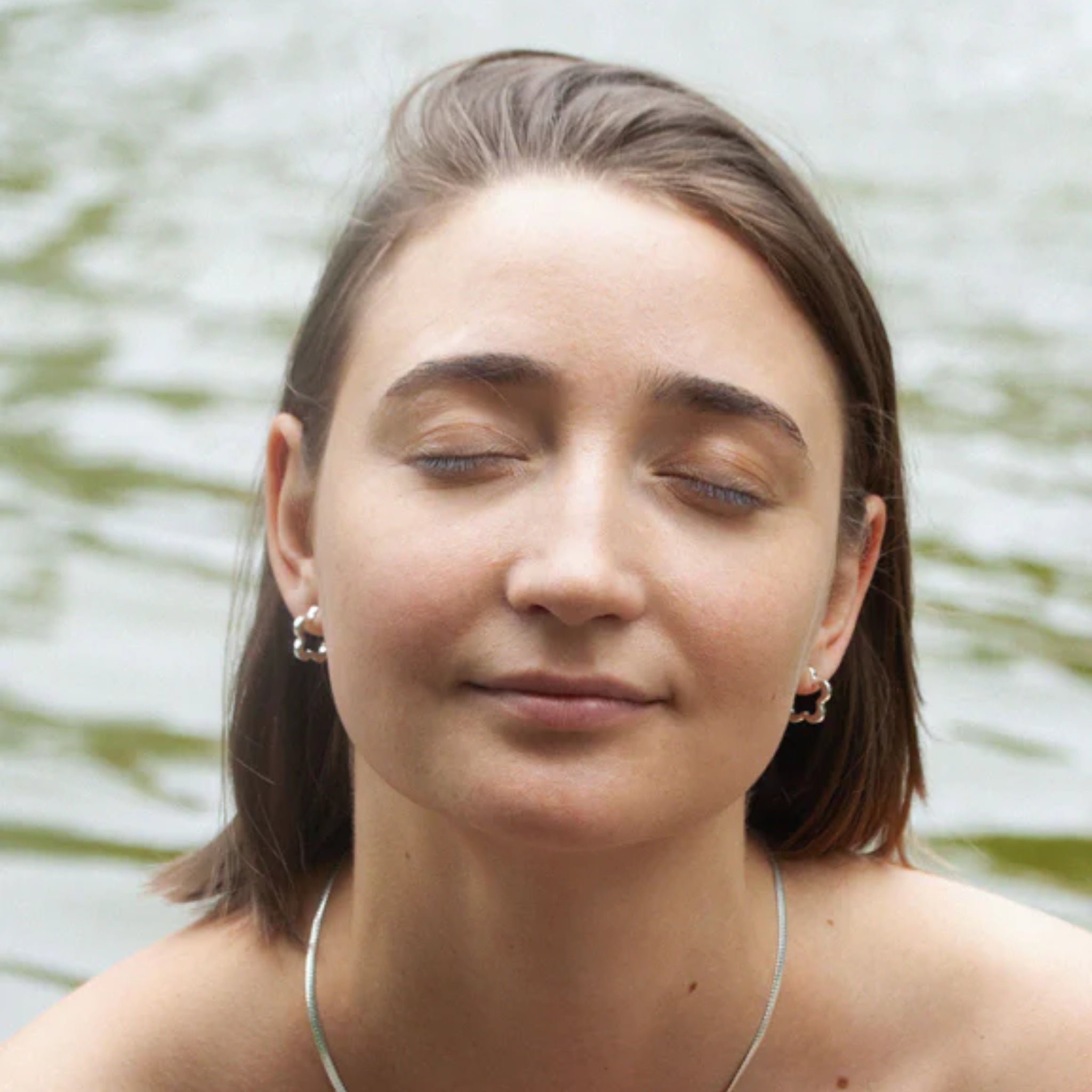 Frau mit geschlossenen Augen und Ohrringen steht vor einem verschwommenen Hintergrund aus Wasser.