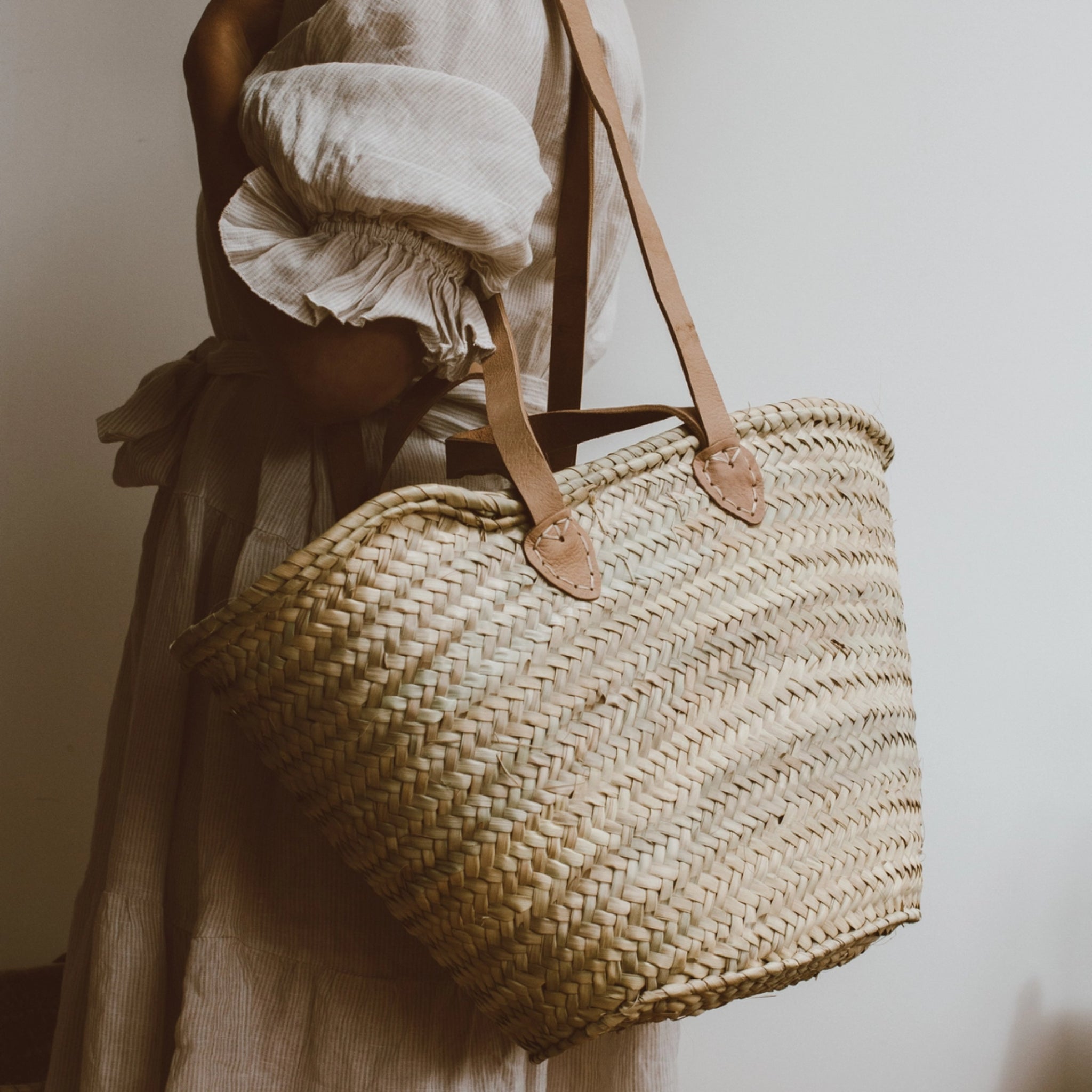 Person in a beige dress holding a large woven straw bag with leather handles against a plain background.