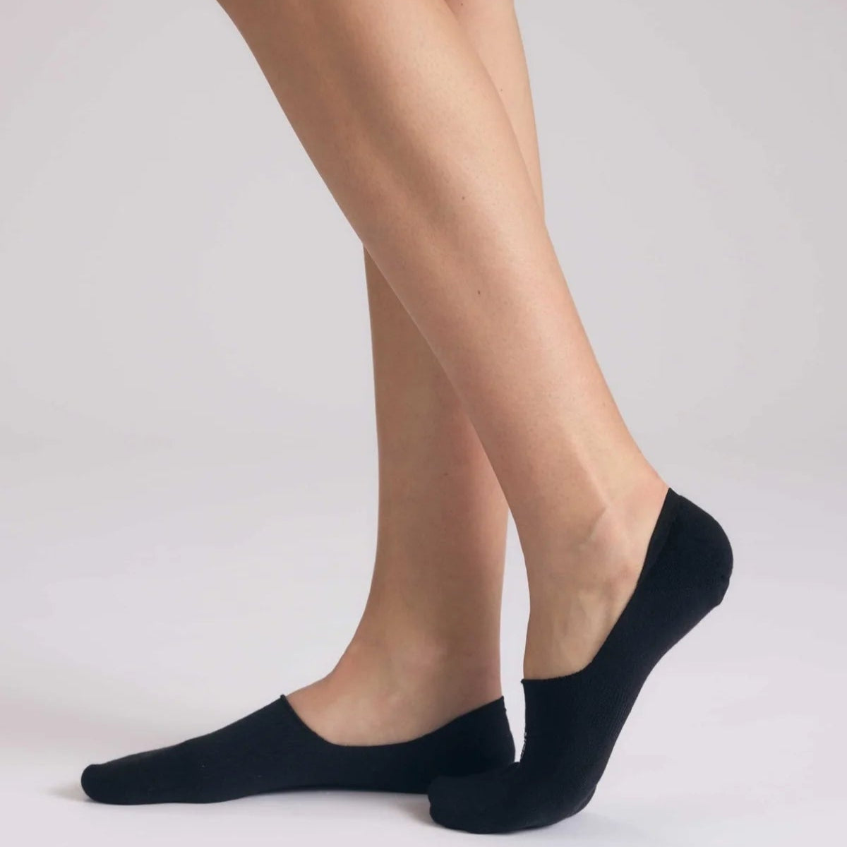 Eine Person, die schwarze No-Show-Socken trägt, steht mit einem flachen Fuß auf einer hellen Unterlage, während der andere auf den Zehenspitzen angehoben ist, so dass die Socken nur die Füße bedecken.