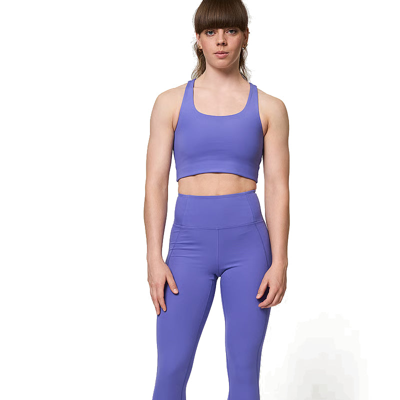 Eine Person steht mit dem Gesicht nach vorne, trägt einen passenden lila Sport-BH und hoch taillierte Leggings und hat einen neutralen Gesichtsausdruck vor einem schlichten weißen Hintergrund.