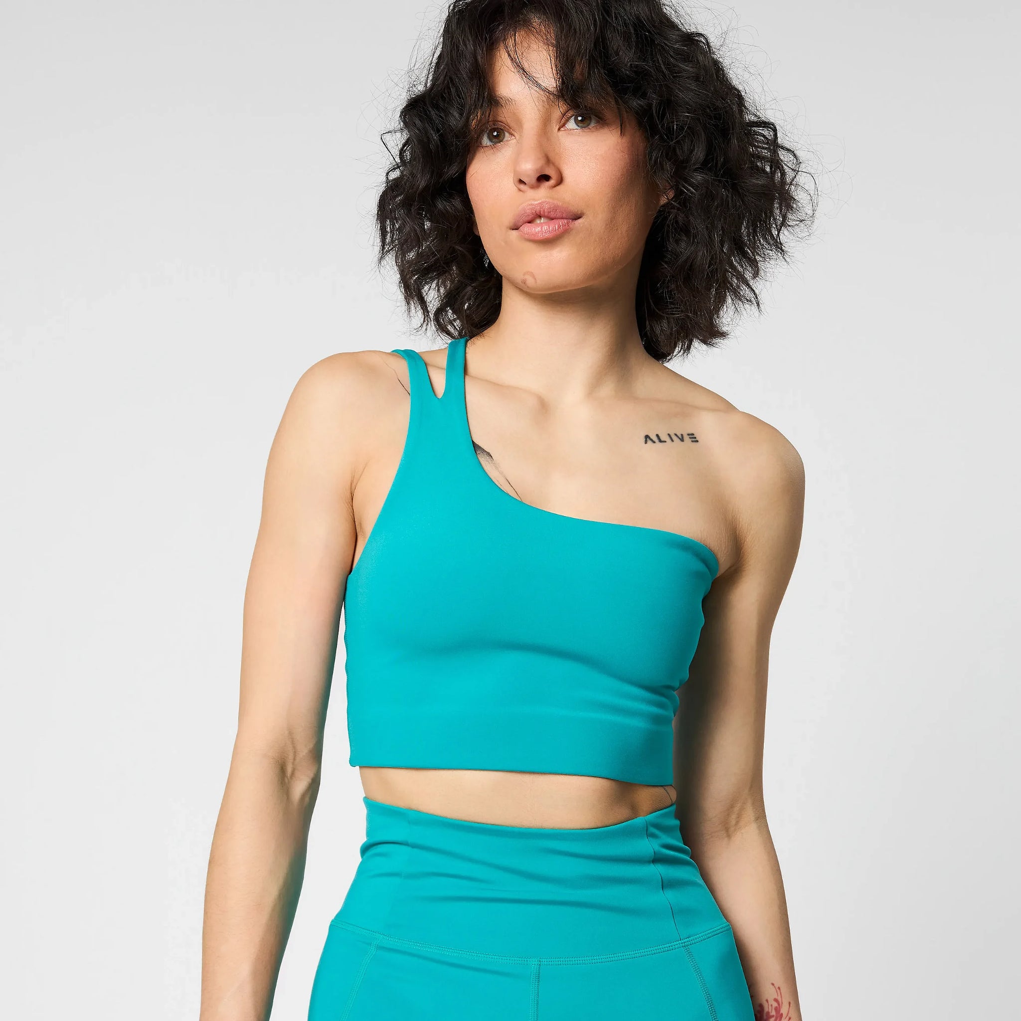 Eine Frau mit lockigem, dunklem Haar trägt ein blaugrünes, schulterfreies Crop-Top und eine dazu passende, hoch taillierte Hose. Sie hat eine Tätowierung mit dem Wort ALIVE auf ihrem Schlüsselbein und blickt leicht zur Seite vor einem schlichten hellen Hintergrund.