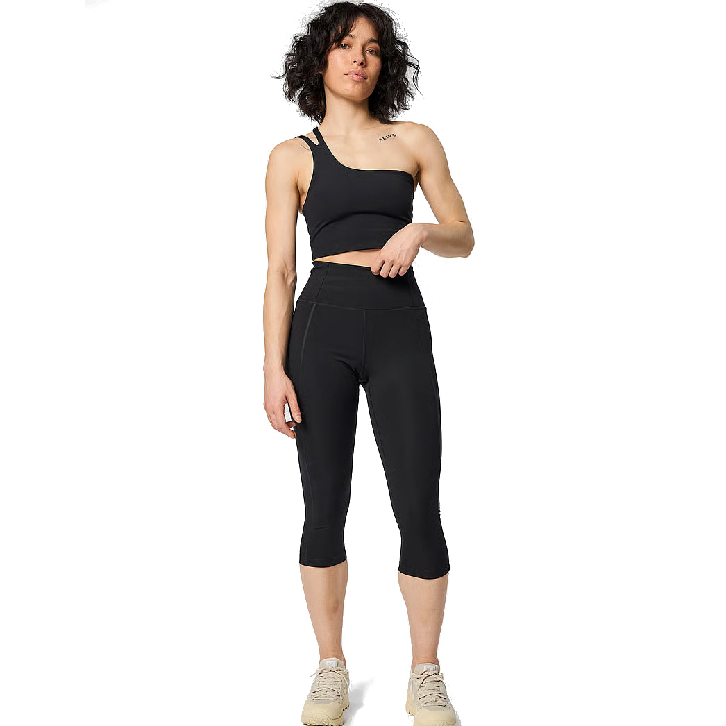 Eine Person mit lockigem, dunklem Haar trägt ein schwarzes, schulterfreies Crop-Top, schwarze, hoch taillierte Capri-Leggings und beigefarbene Turnschuhe. Sie steht mit entspannter Körperhaltung vor einem weißen Hintergrund.