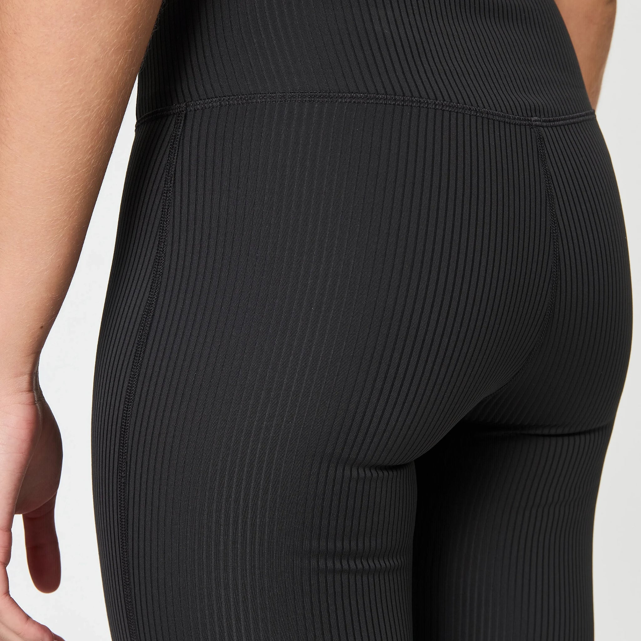 Nahaufnahme einer Person, die schwarze, gerippte, figurbetonte Leggings trägt und ihre Hand vor einem unifarbenen Hintergrund an die Seite legt.