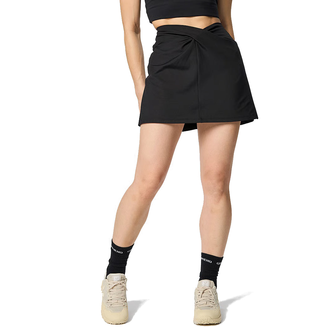 Eine Person, die einen schwarzen Minirock mit verdrehter Front, ein schwarzes Crop-Top, schwarze Socken und cremefarbene Turnschuhe trägt und vor einem weißen Hintergrund steht.