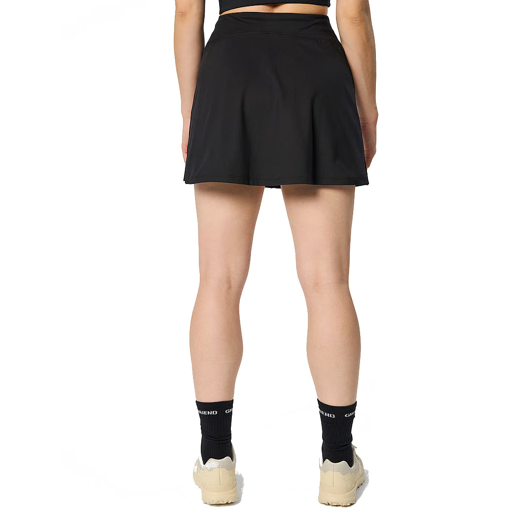 Eine Person steht mit dem Gesicht nach unten und trägt einen schwarzen Sportrock, schwarze Socken, beigefarbene Turnschuhe und ein schwarzes Oberteil. Der Hintergrund ist einfarbig weiß.