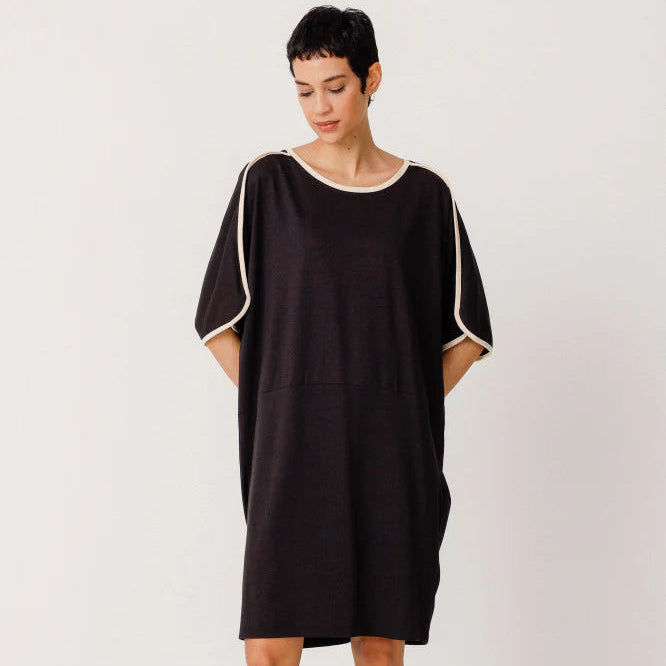 Eine Person mit kurzen dunklen Haaren trägt ein locker sitzendes schwarzes Kleid mit weißer Borte an den Ärmeln und am Halsausschnitt, dazu kniehohe schwarze Stiefel, die vor einem schlichten weißen Hintergrund stehen.